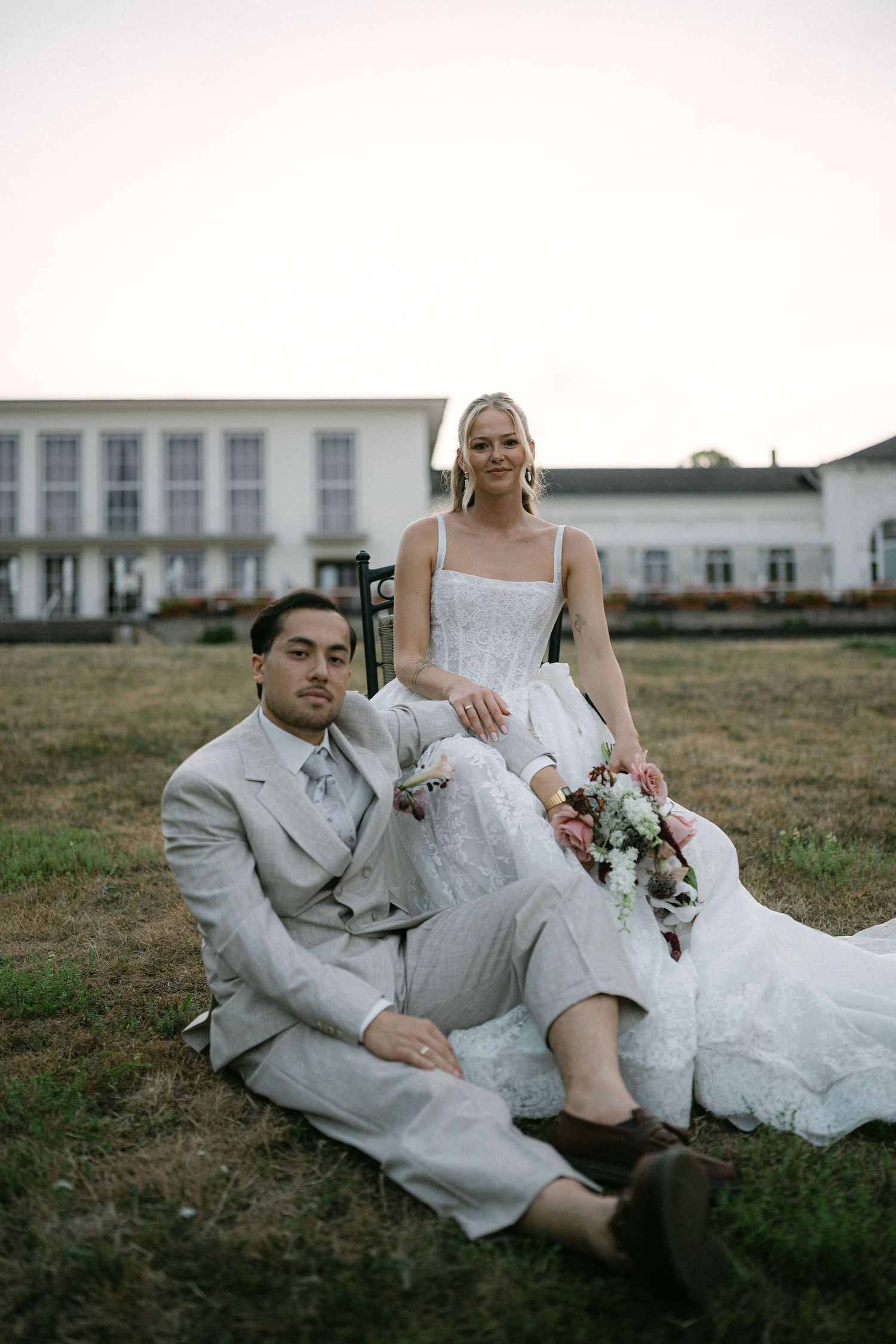 Bride in white gown holds bouquet seated behind groom in light gray suit on grass before elegant white venue.
