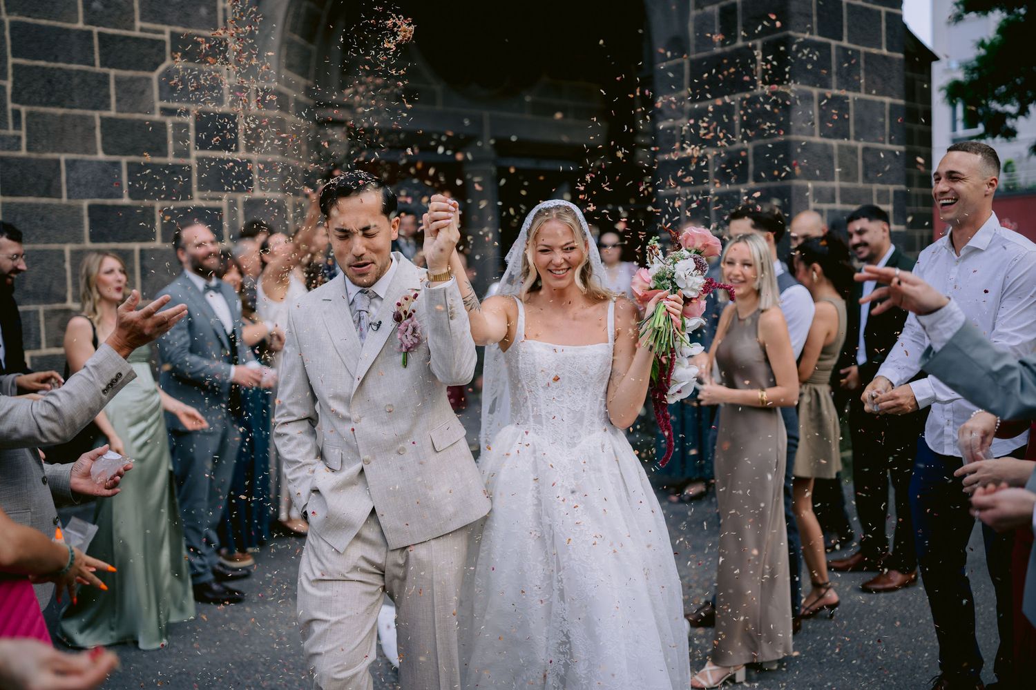 Bride and groom walk through confetti shower outside stone church surrounded by cheering wedding guests.