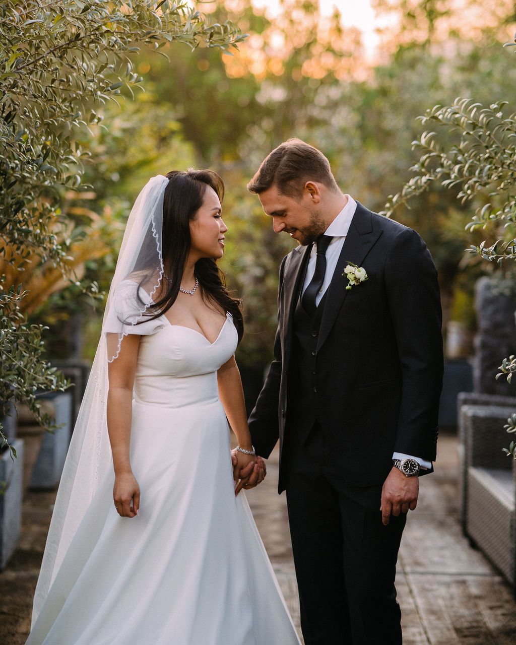 A couple in wedding attire share a romantic moment on a garden path surrounded by lush foliage at sunset.