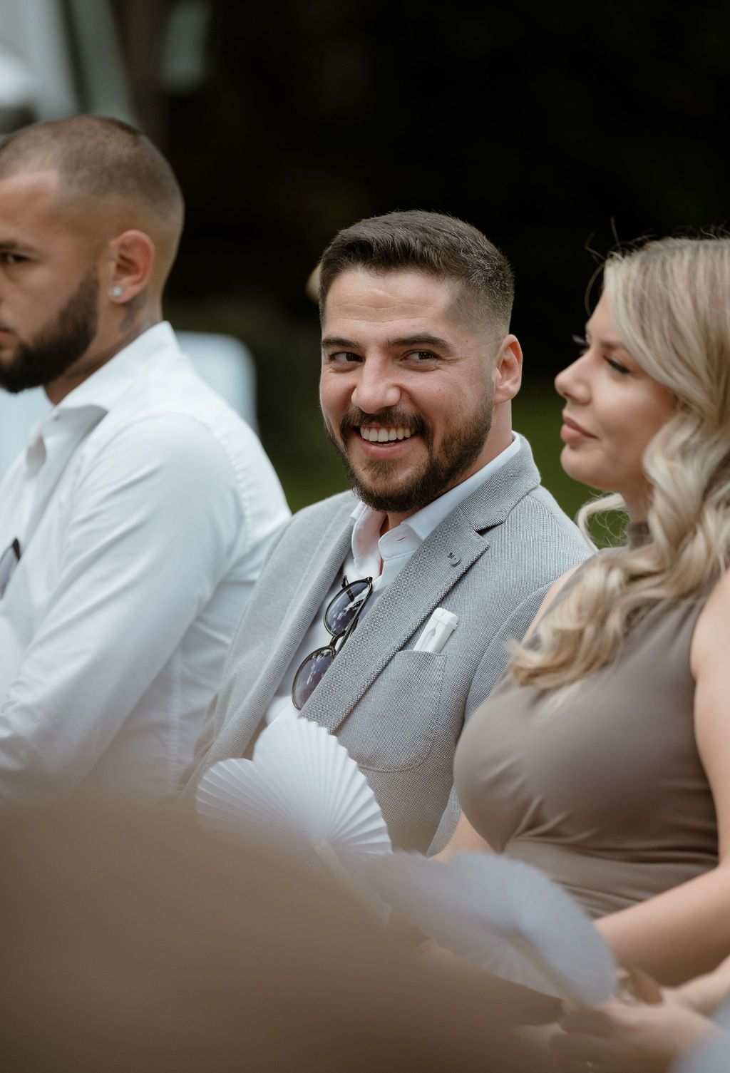 Wedding guests dressed in grey suit and formal attire smile during outdoor ceremony while holding hand fans.