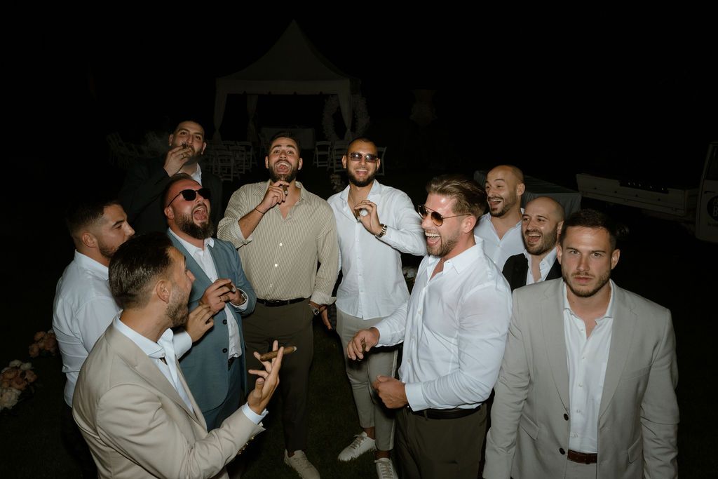 Groomsmen and guests enjoying themselves at a wedding reception in formal attire against a dark background.