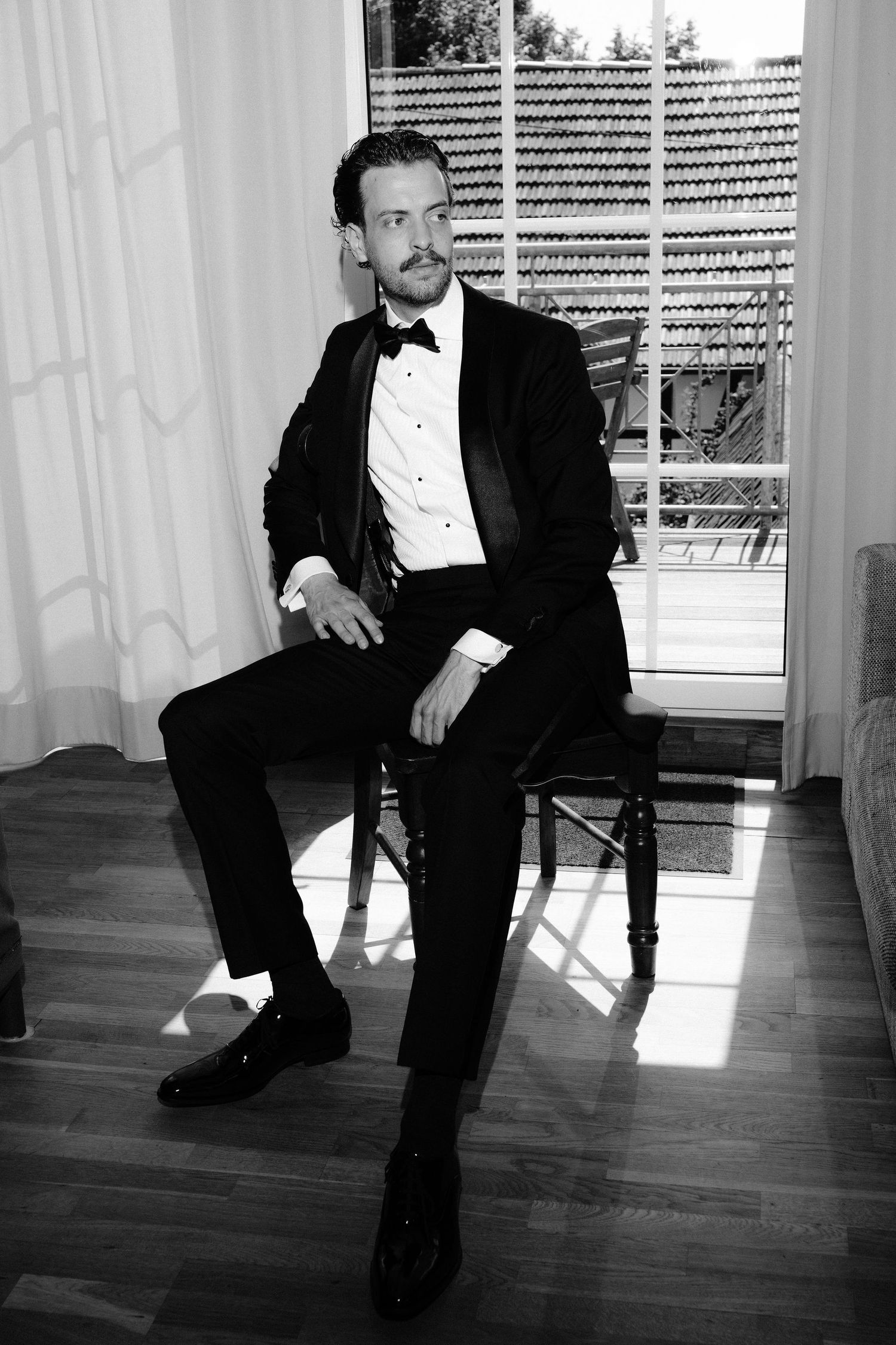Man in black tuxedo with bow tie sitting on chair in black and white portrait photo.