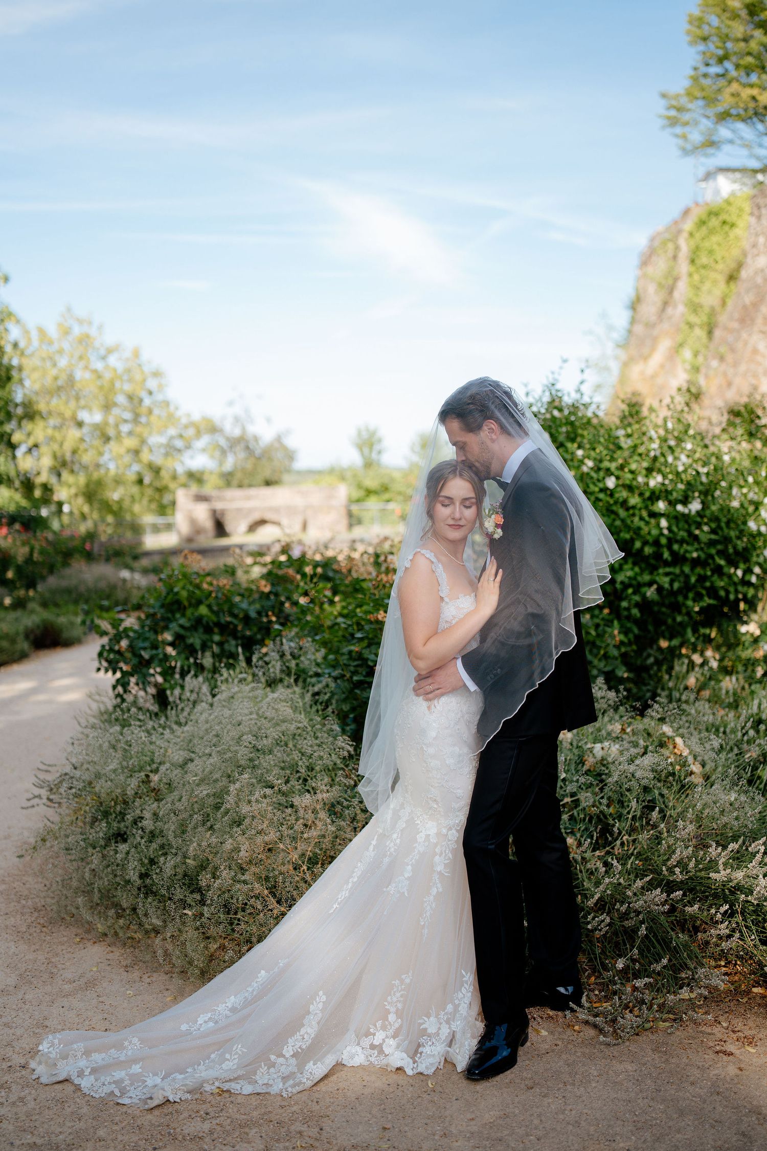 Bride and groom embrace under veil in lush garden, wearing elegant lace wedding gown with train and black suit.