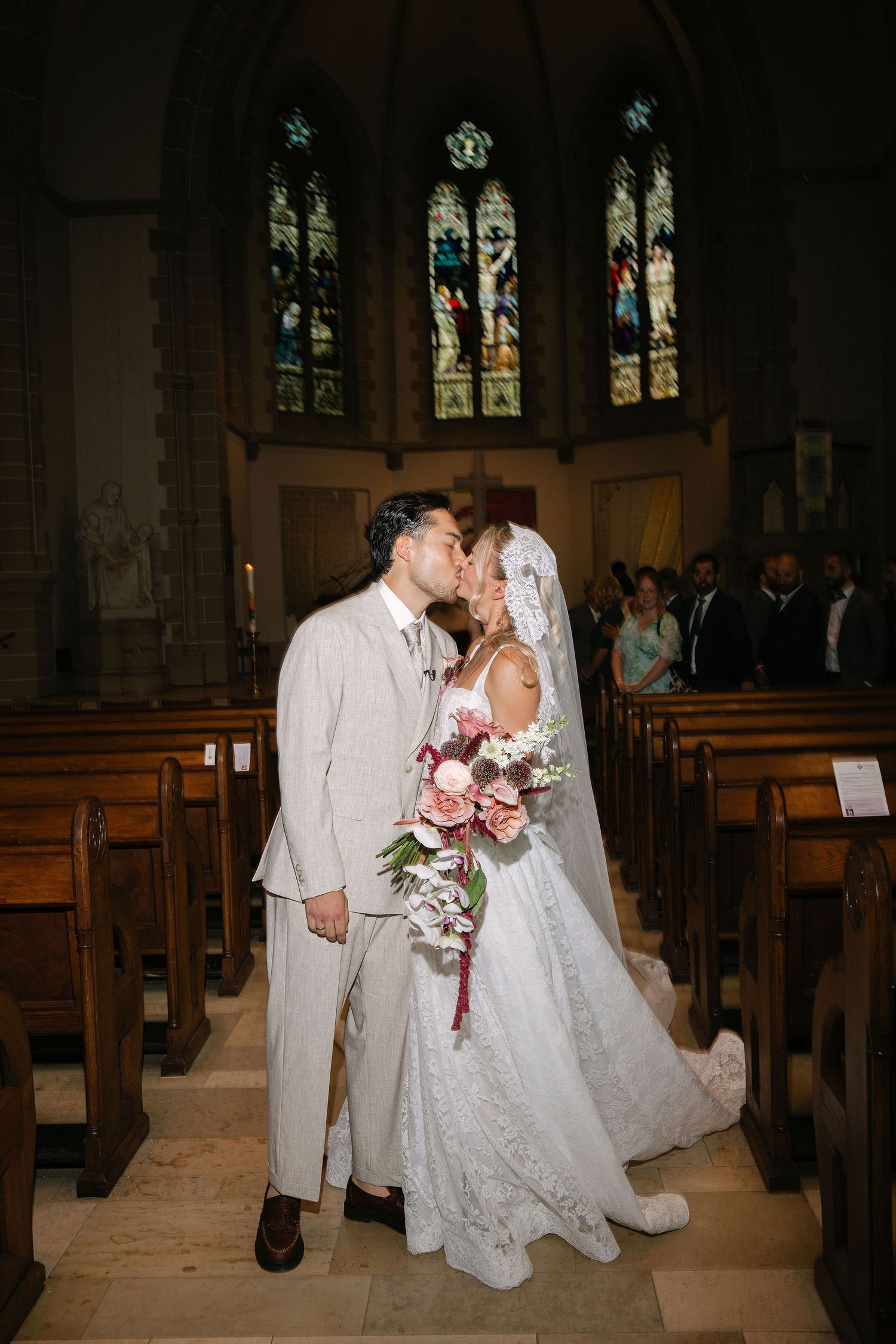 Newlyweds share first kiss in ornate church aisle with stained glass windows and wooden pews.