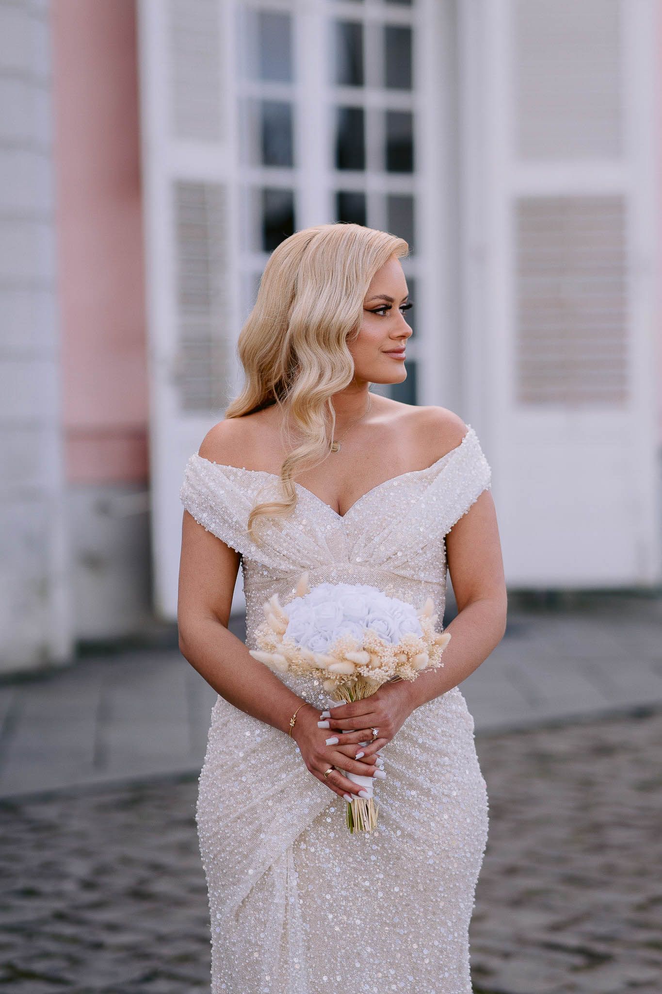 Beautiful bride in off-shoulder sequined wedding dress holds white bouquet while standing outside elegant building.