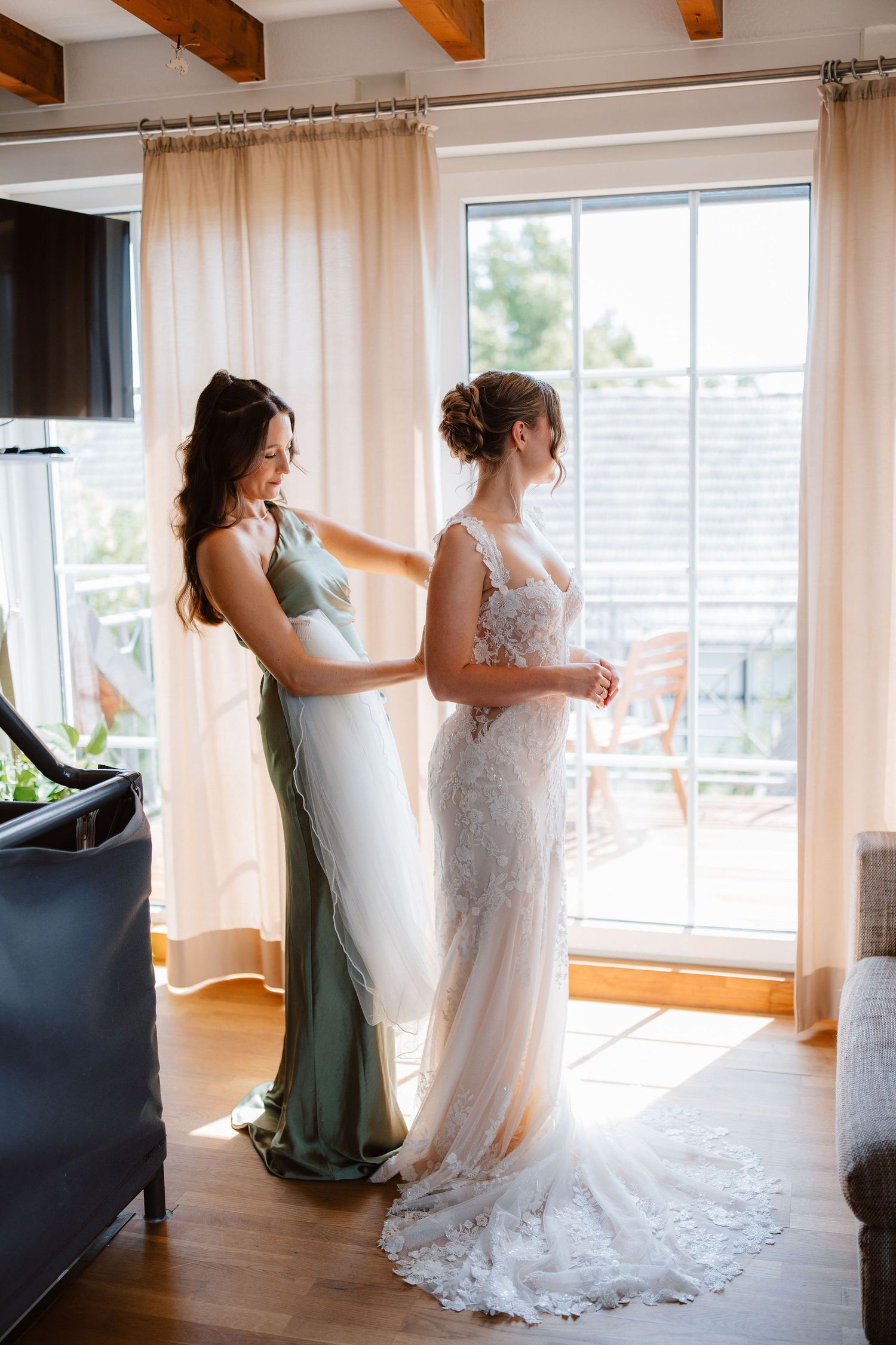 Bridesmaid in sage green dress buttoning the back of brides white lace wedding gown by window.