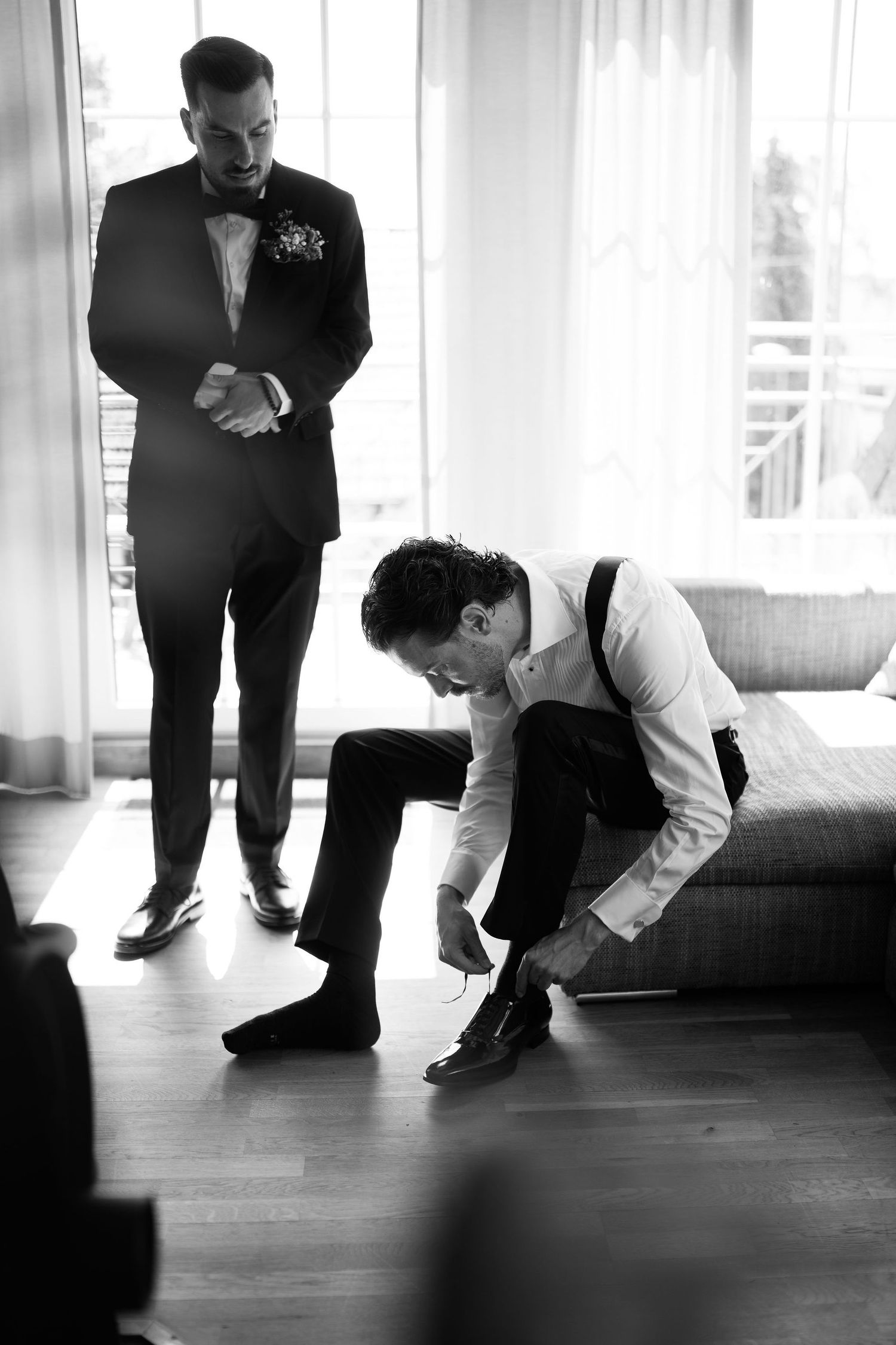 Black and white photo of groom tying his shoes while groomsman watches in a sunlit hotel room.