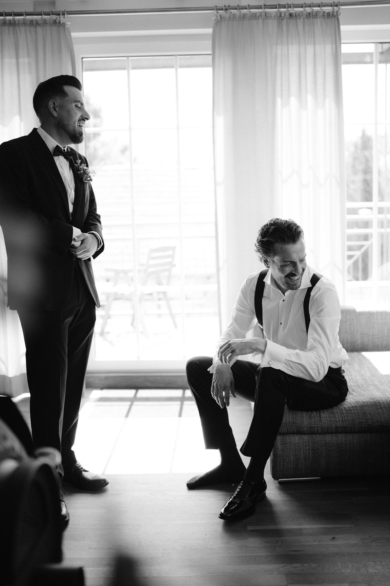 Black and white photo of a groom in a tuxedo laughing with his groomsman while getting ready for the wedding.