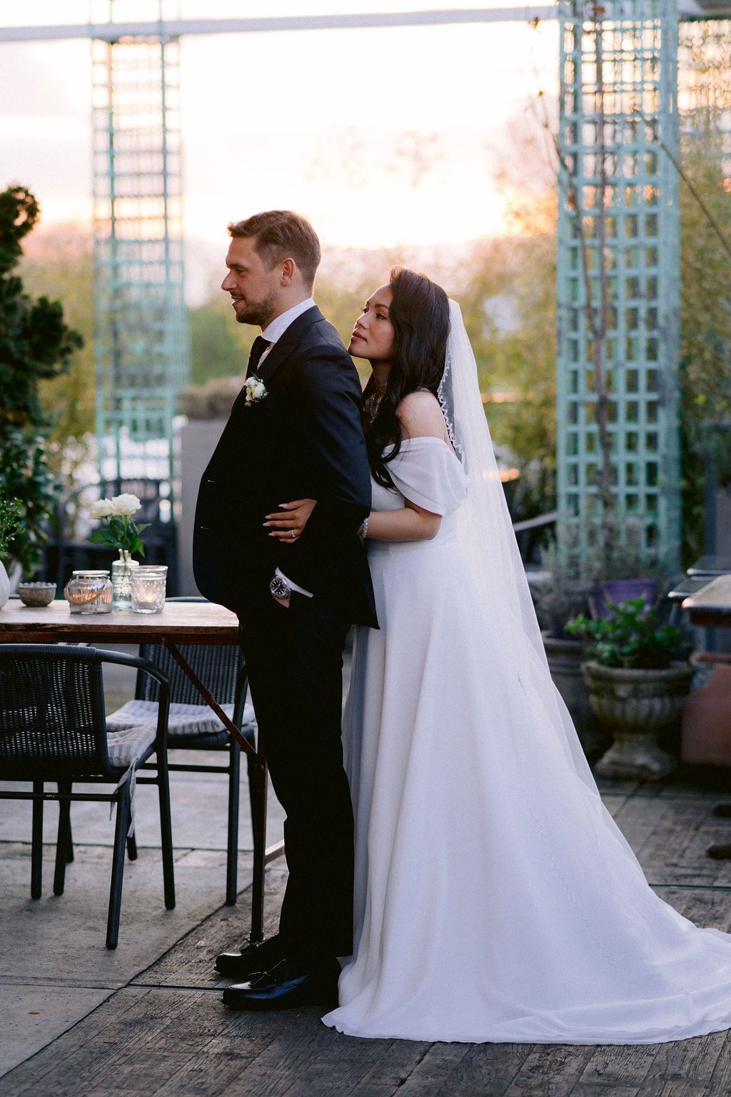 A couple embraces during their wedding reception on an outdoor patio with garden views at sunset.