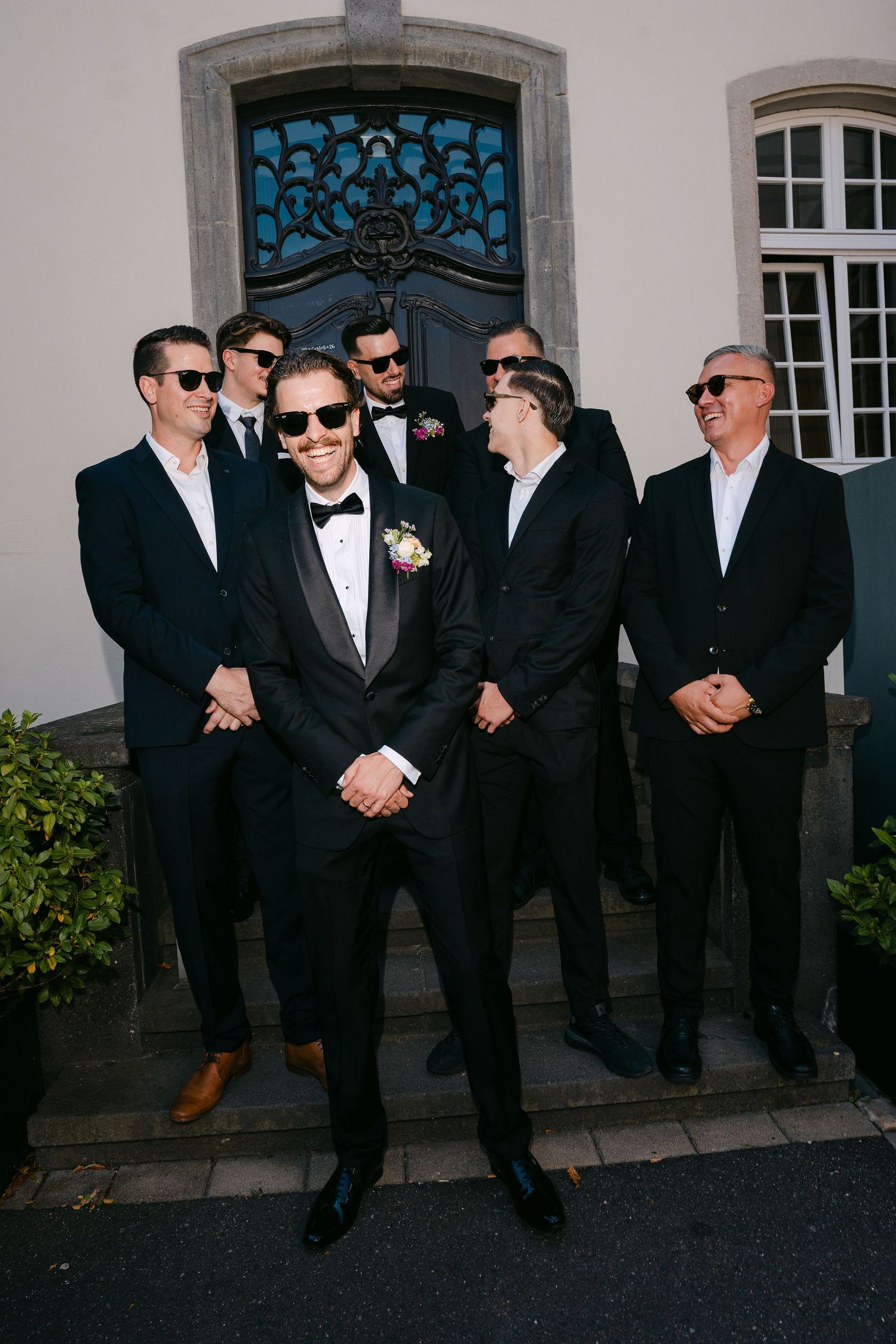 Groom in black tuxedo poses with groomsmen in suits and sunglasses on venue steps.