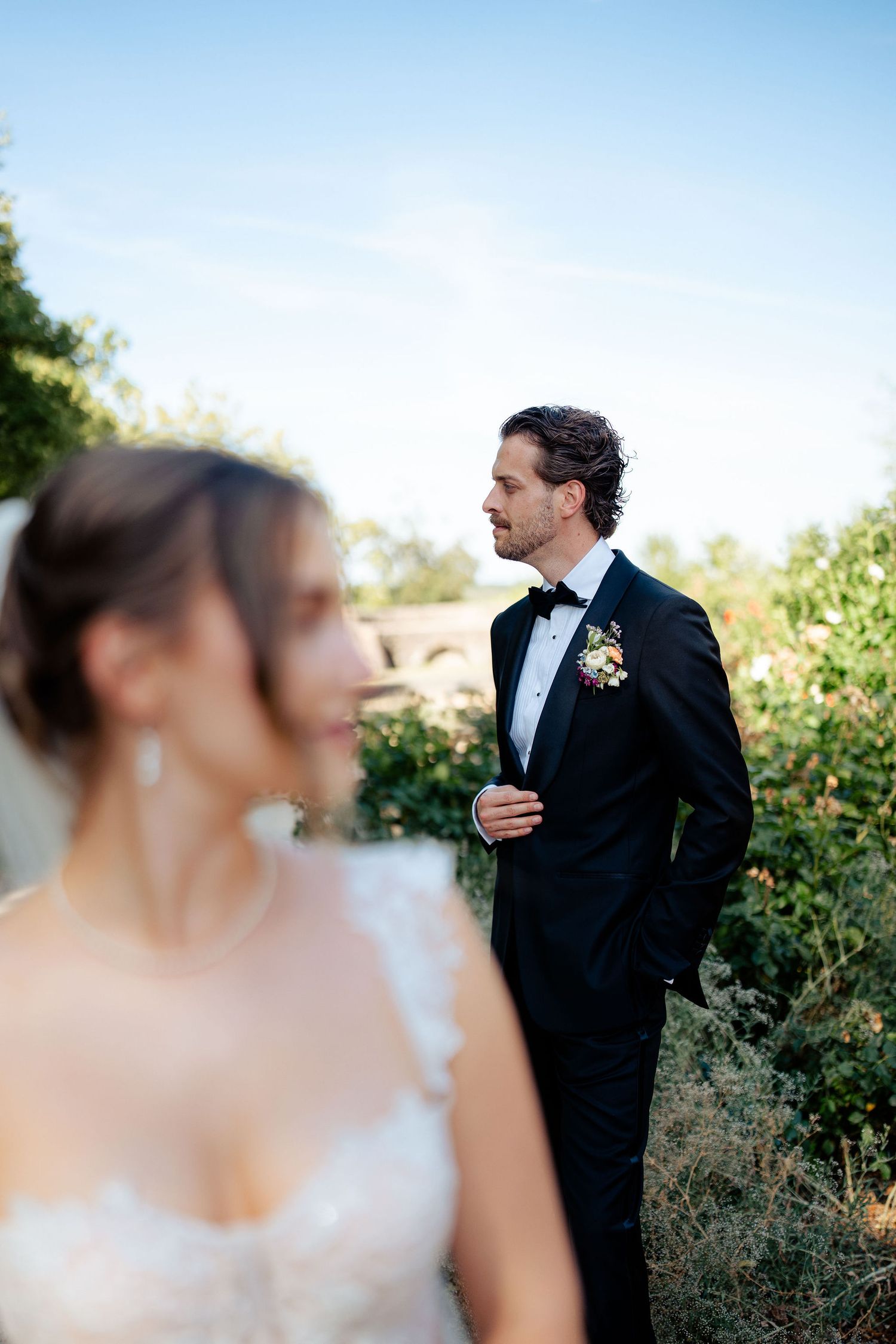 Groom in black tuxedo with boutonniere stands outdoors, bride in white dress blurred in foreground.
