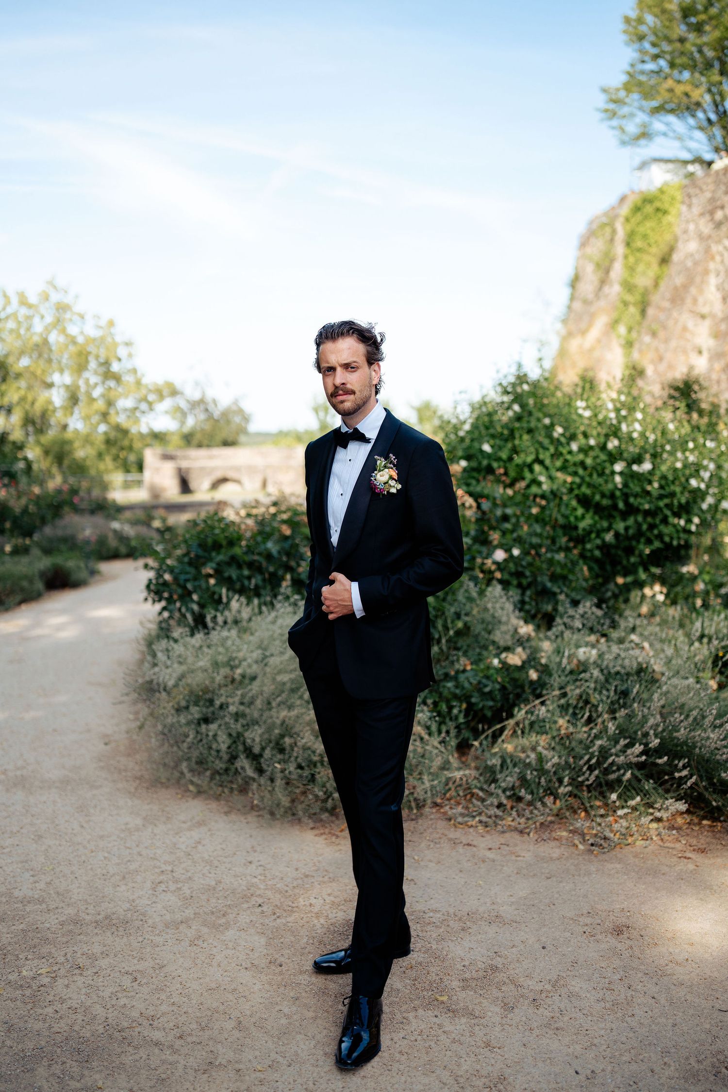 Groom in black tuxedo with boutonniere poses on garden path at outdoor wedding venue.