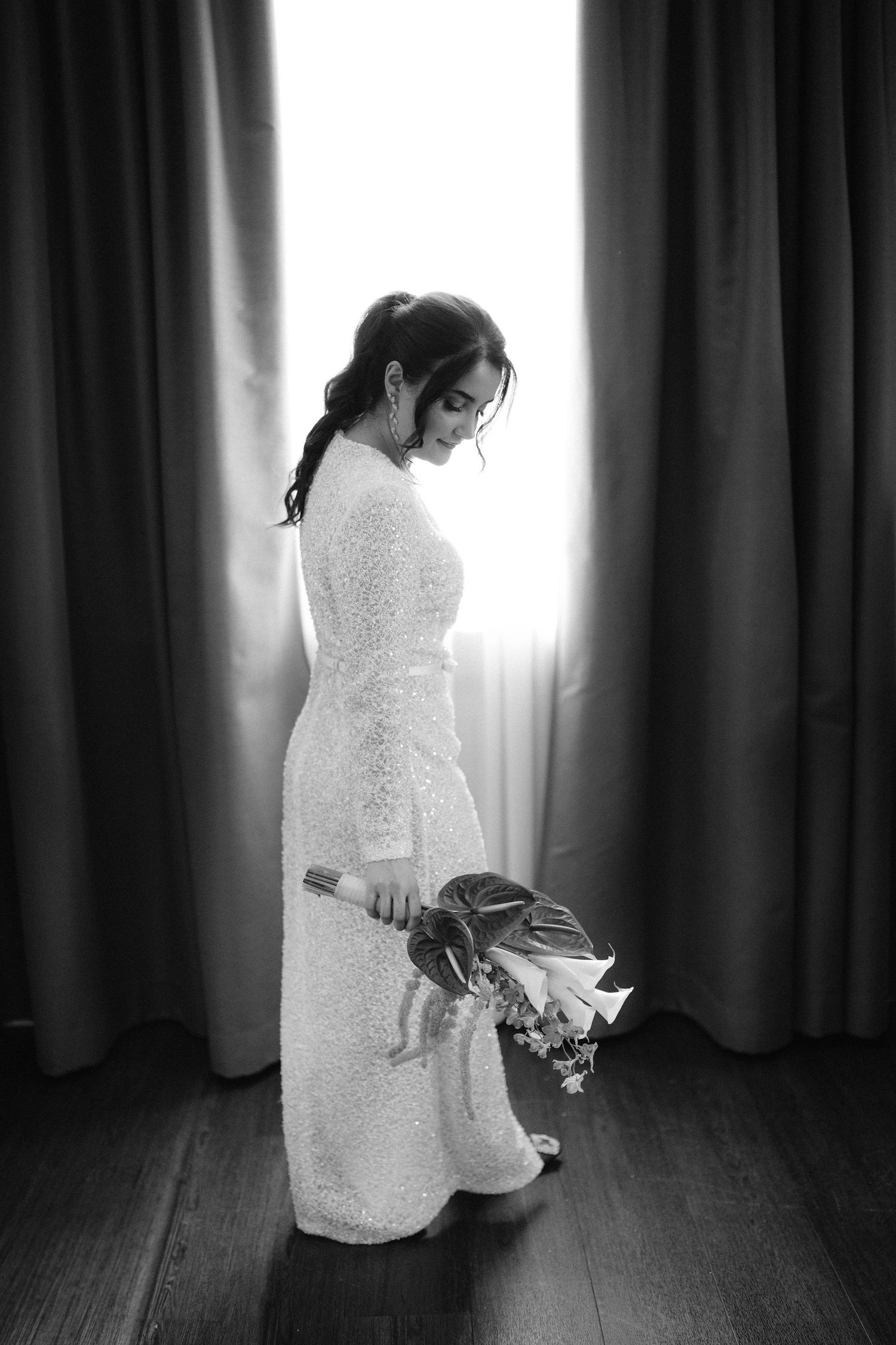 Black and white photo of a bride in a lace gown holding a bouquet, standing by curtained window.