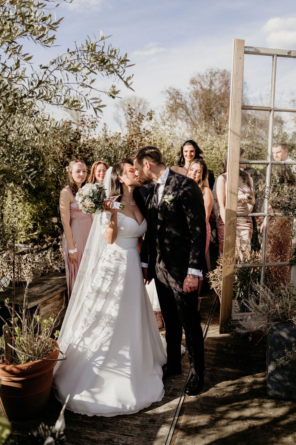 A romantic outdoor wedding ceremony with a white-gowned bride and groom beneath trees on a sunny afternoon.