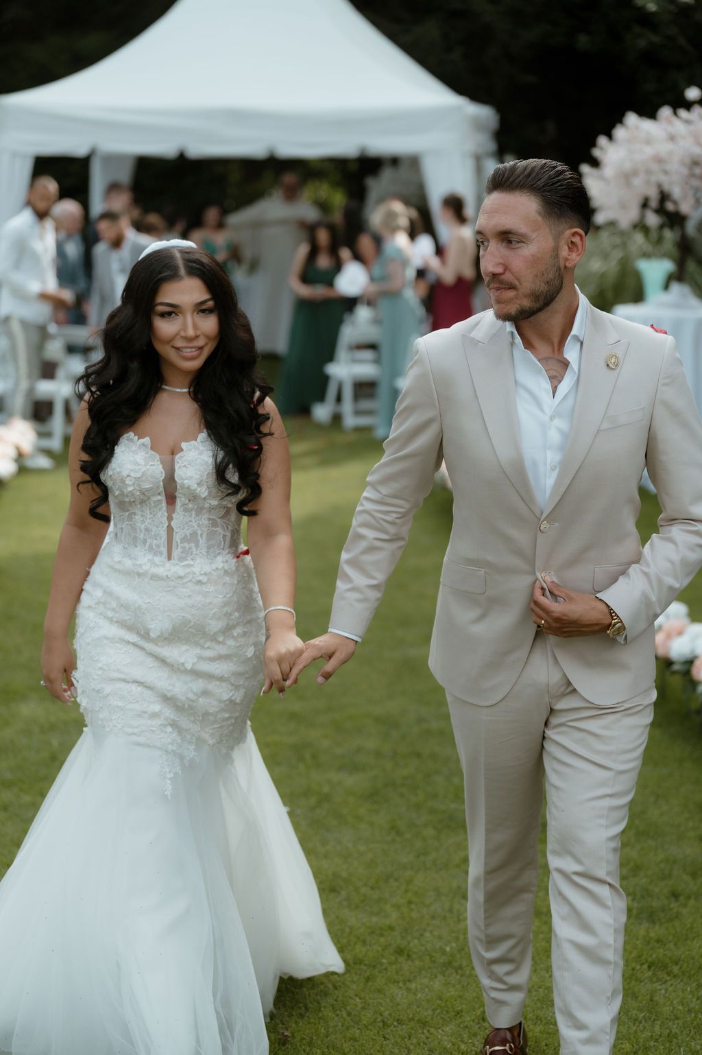 A couple in wedding attire walks together on a lush green lawn during an outdoor celebration.