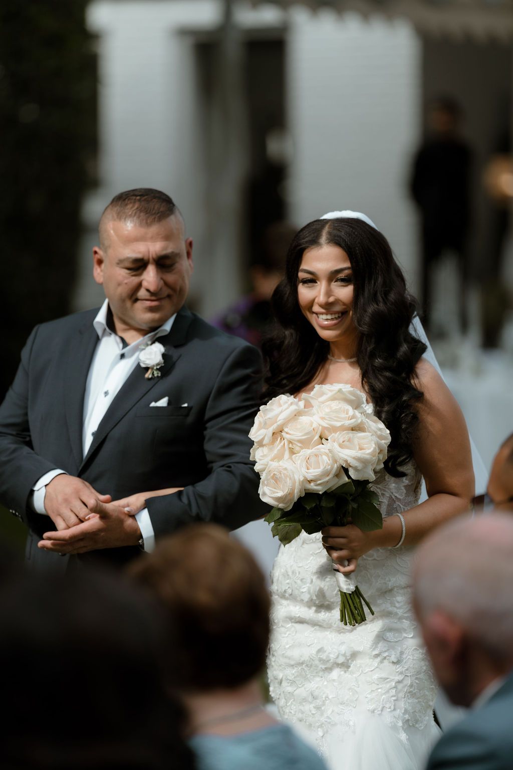 A bride in a lace wedding dress holds a white rose bouquet while smiling during an outdoor wedding ceremony.