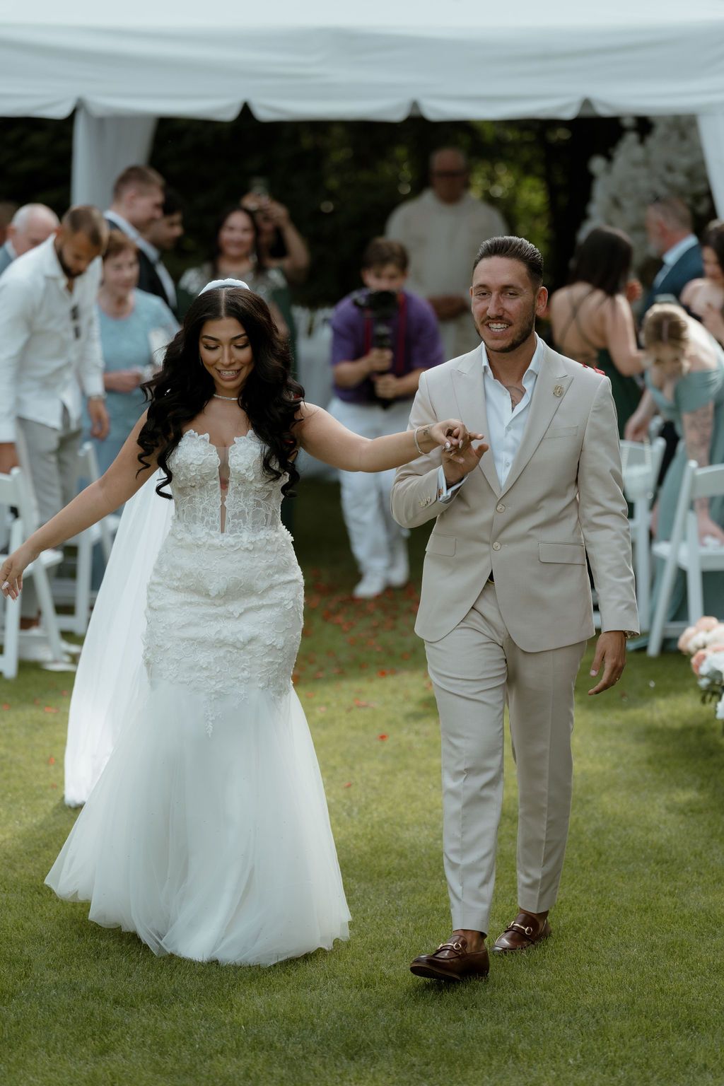 Happy newlyweds celebrate during an outdoor wedding ceremony, with the bride in white and groom in beige walking on grass.