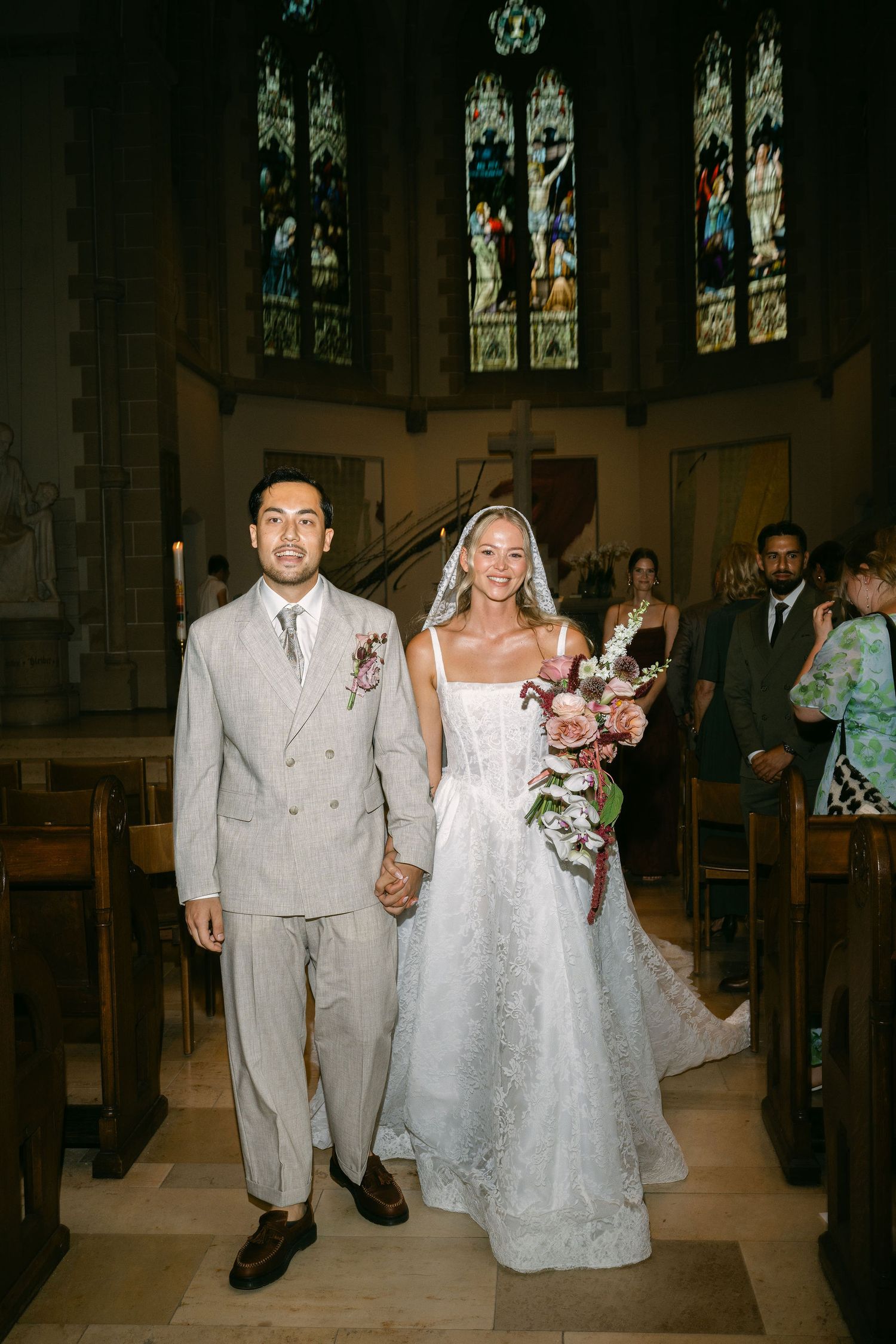 Newlywed couple walks down church aisle, bride in lace gown with bouquet, groom in grey suit, stained glass windows behind.