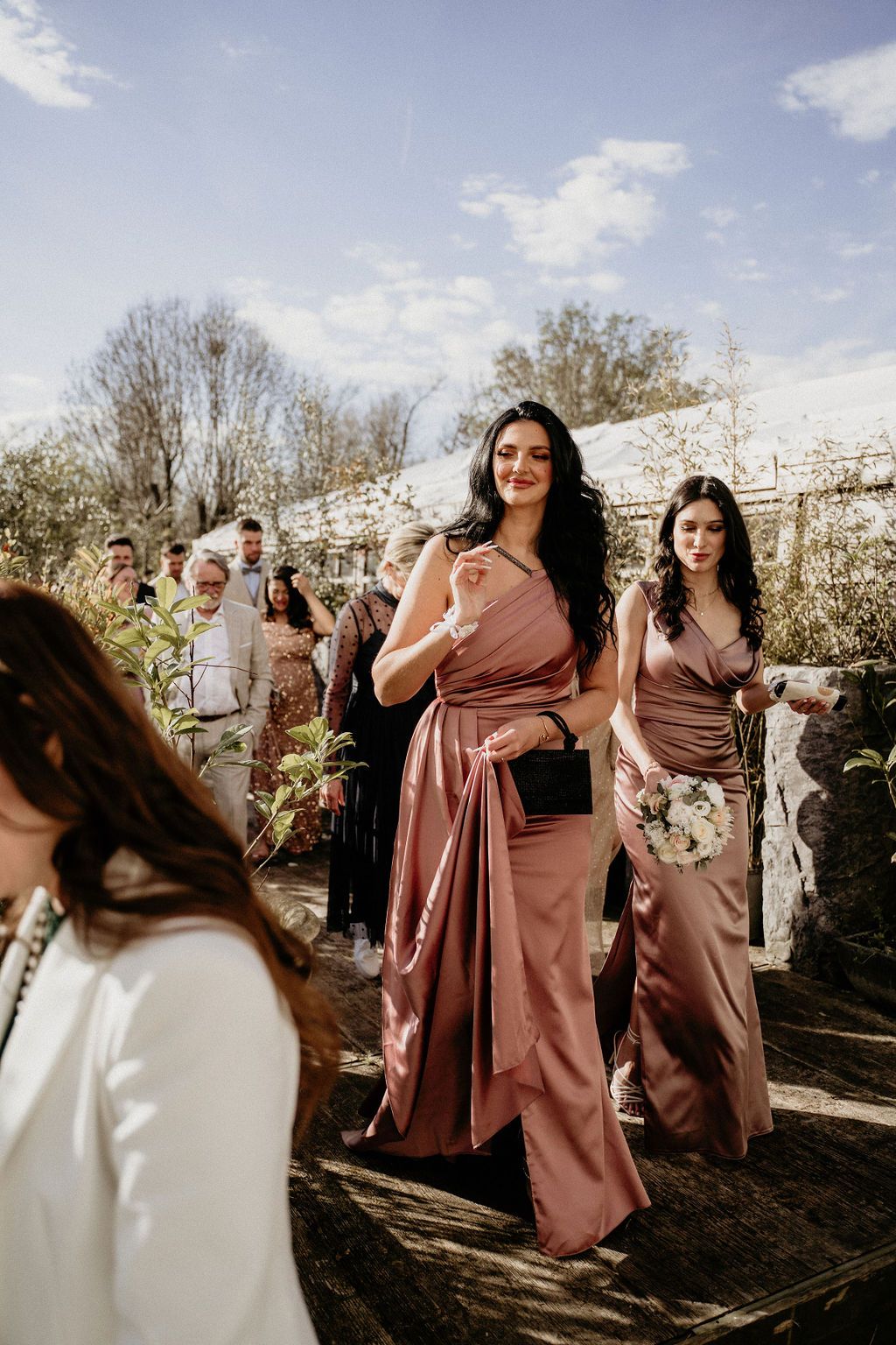 Joyful wedding party wearing dusty rose bridesmaid dresses dance and celebrate outdoors on a sunny afternoon.