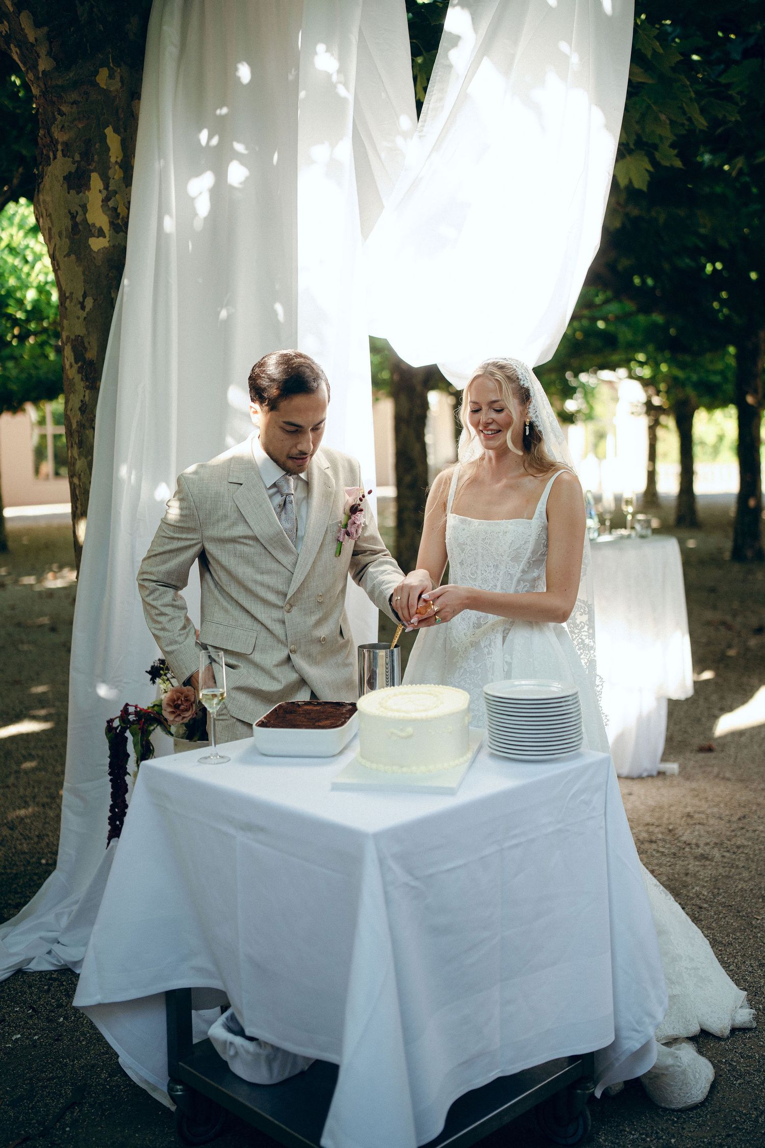 Smiling bride and groom cutting their white wedding cake outdoors under elegant white draped canopy.