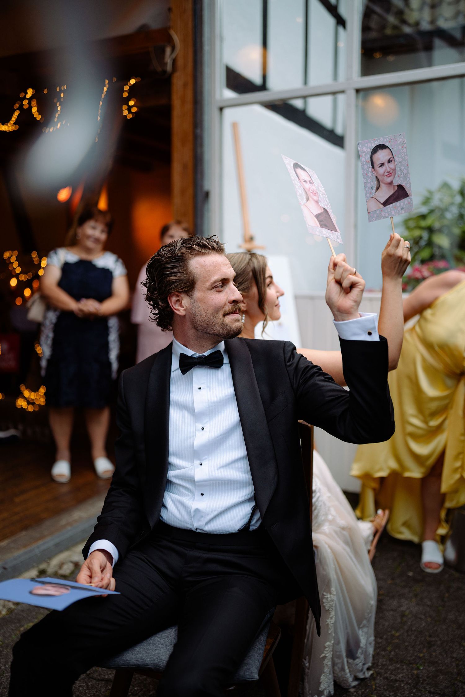 Groom in black tuxedo holds up photo card at wedding reception while bride sits beside him outdoors.
