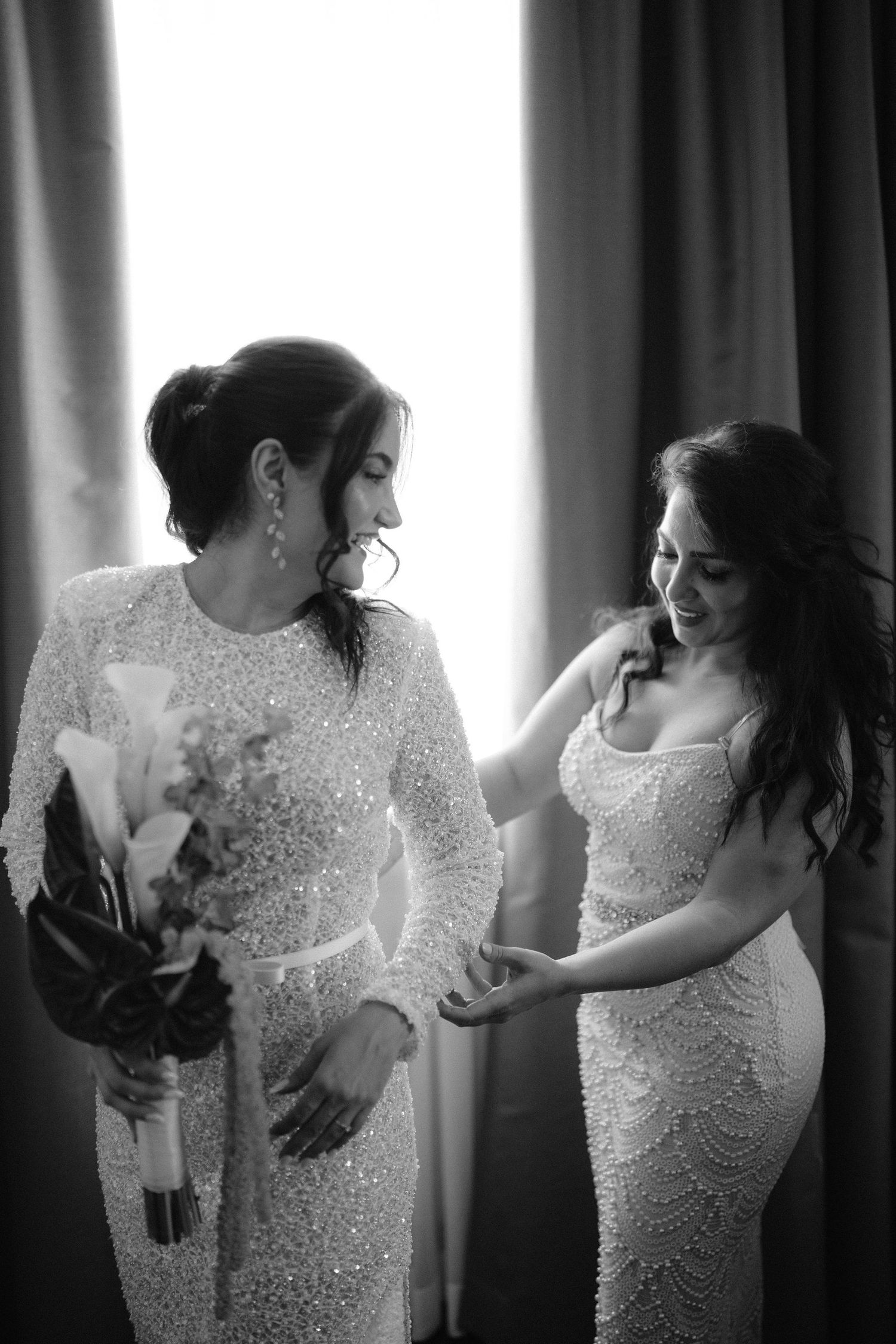 Bride in beaded gown holds bouquet while bridesmaid in sequin dress smiles in black and white photo.