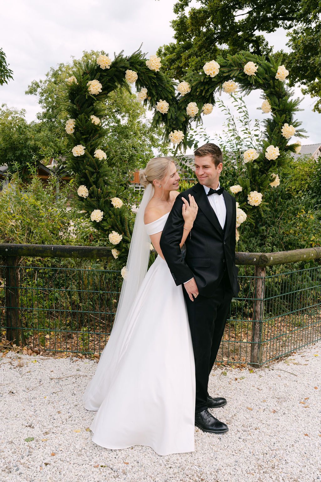 Couple in elegant wedding attire poses beneath climbing roses on a gravel path in a romantic garden setting.