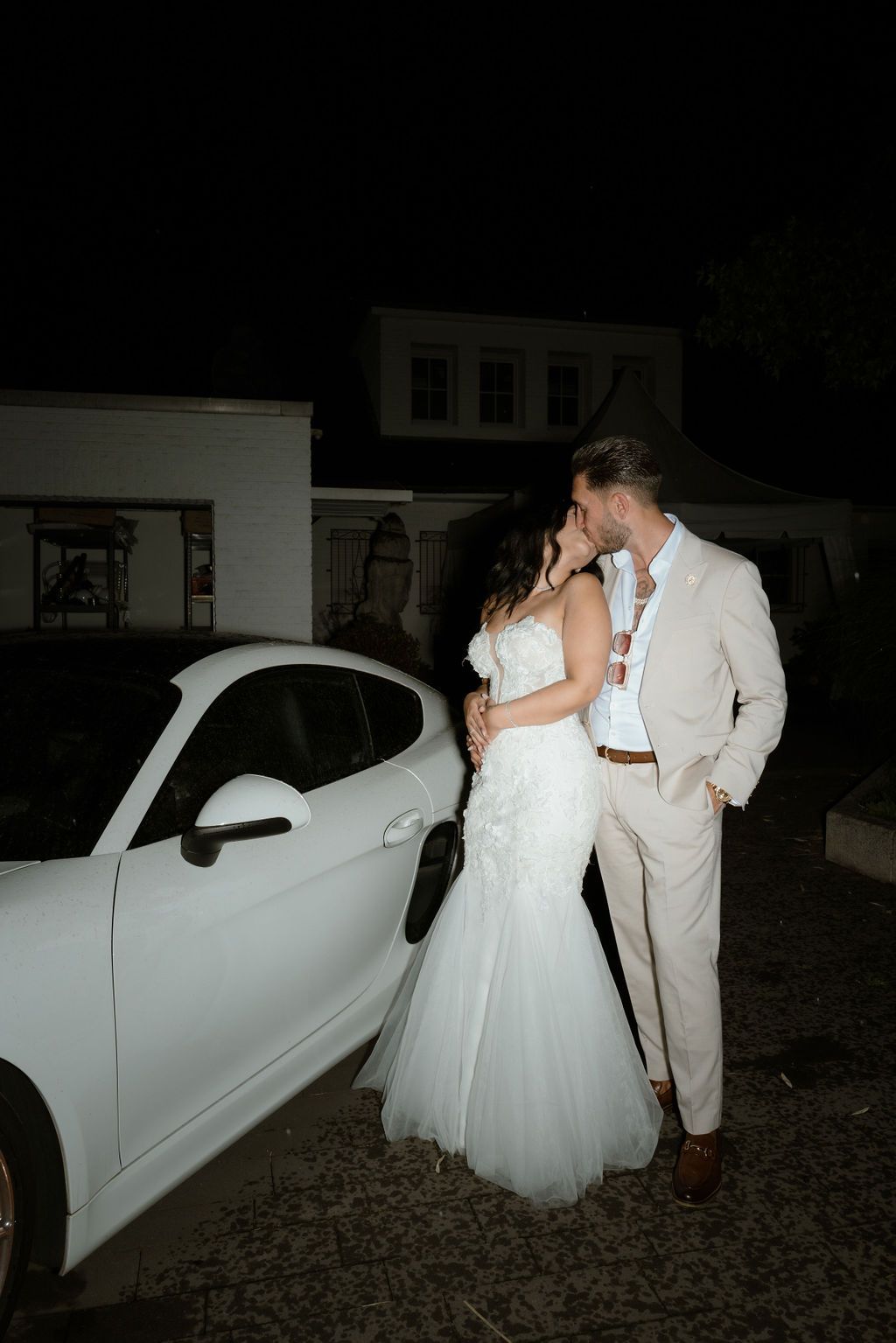 A bride and groom pose for romantic nighttime photos next to a classic white car at their wedding reception.