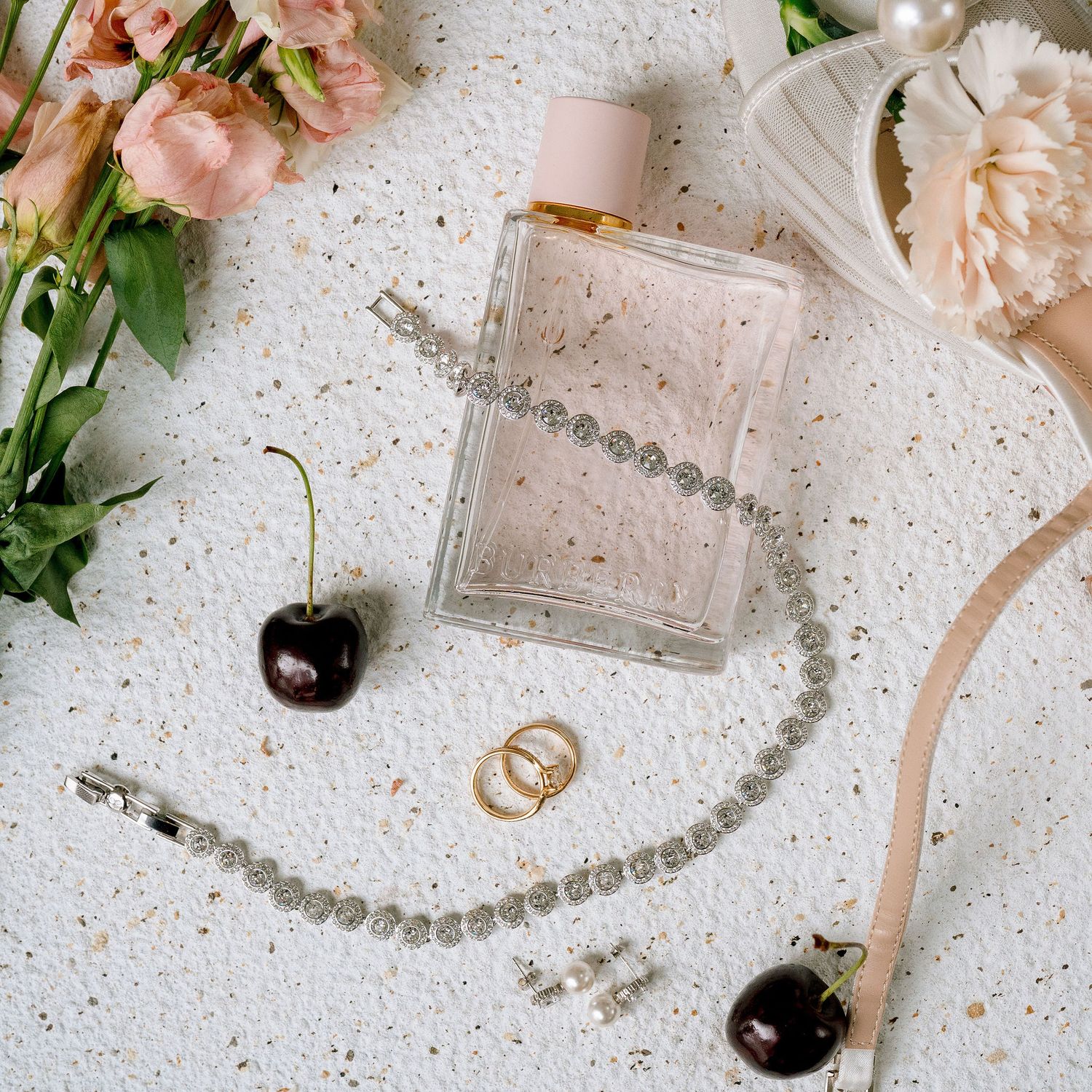 Flat lay of wedding accessories including perfume, diamond bracelet, gold rings, earrings and cherries.