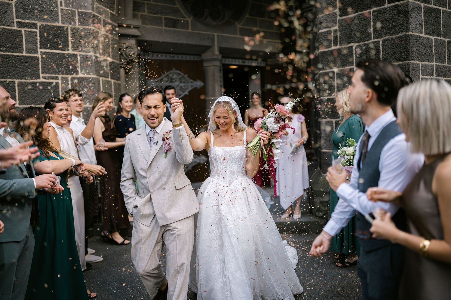 Bride and groom exit church ceremony through cheering guests throwing confetti on their wedding day.