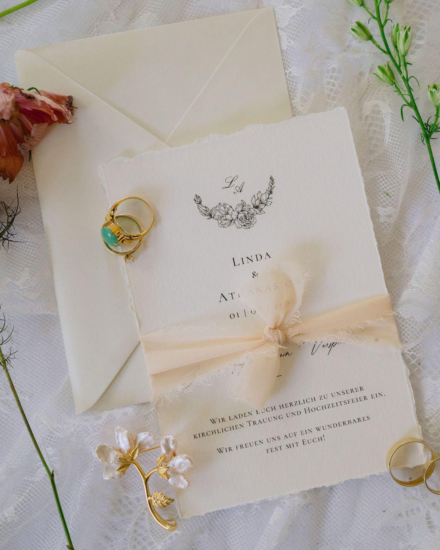 Elegant wedding invitation suite with cream ribbon, gold rings, and floral details on white lace background.