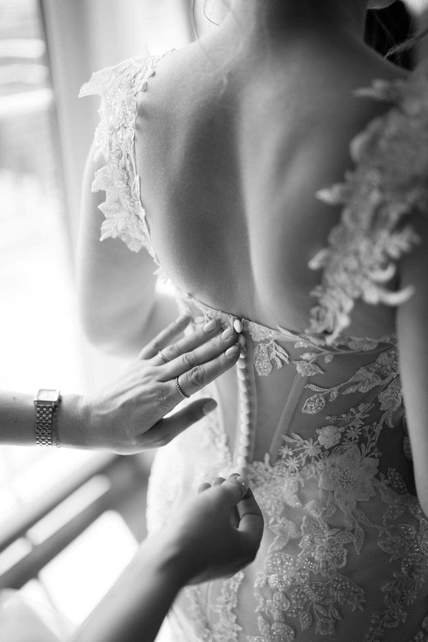 Black and white close-up of hands fastening the intricate lace back buttons of a bridal wedding gown.