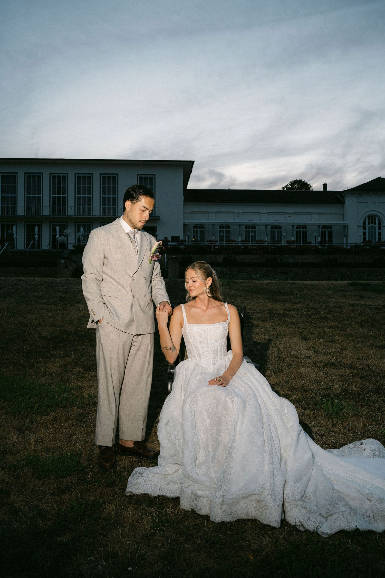 Bride in white lace gown sits outdoors at dusk as groom in beige suit stands beside her near a white building.