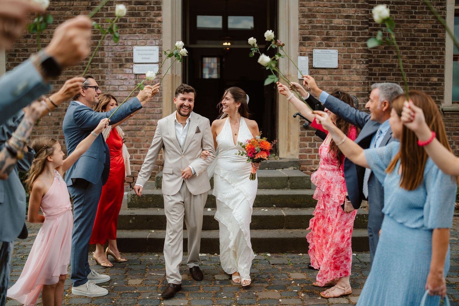 A bride and groom walk through a flower arch created by wedding guests outside a brick church.