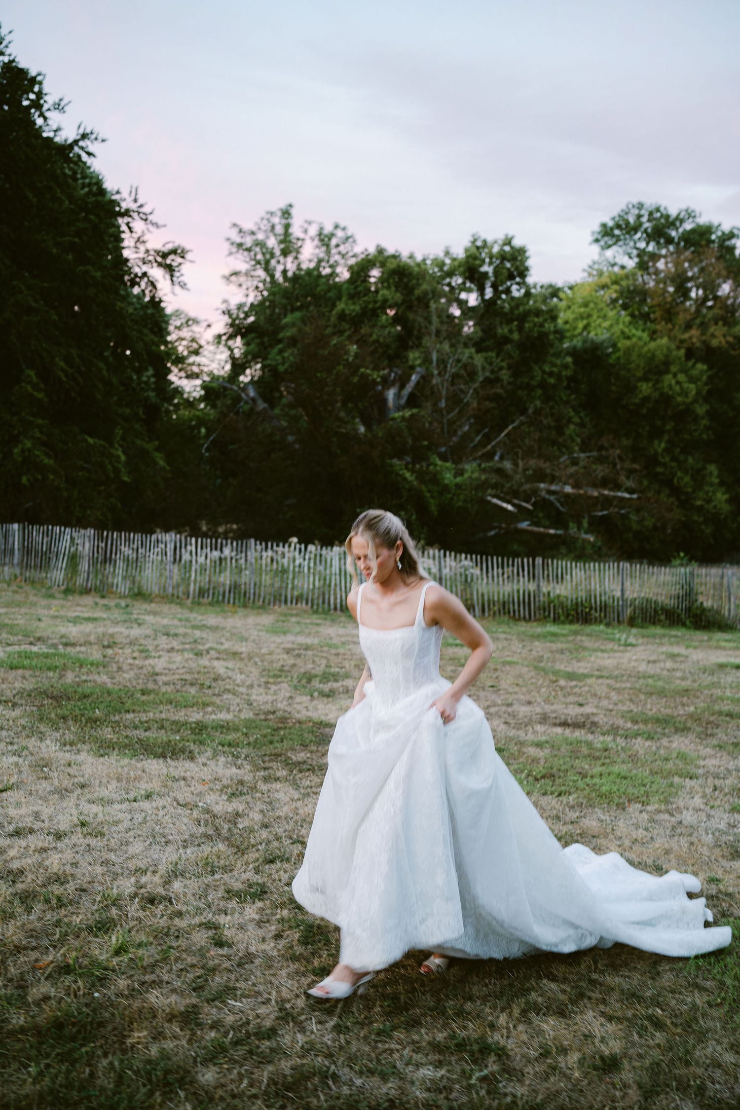 Bride in a flowing white ball gown with train stands on a grassy lawn at dusk, looking down gracefully.