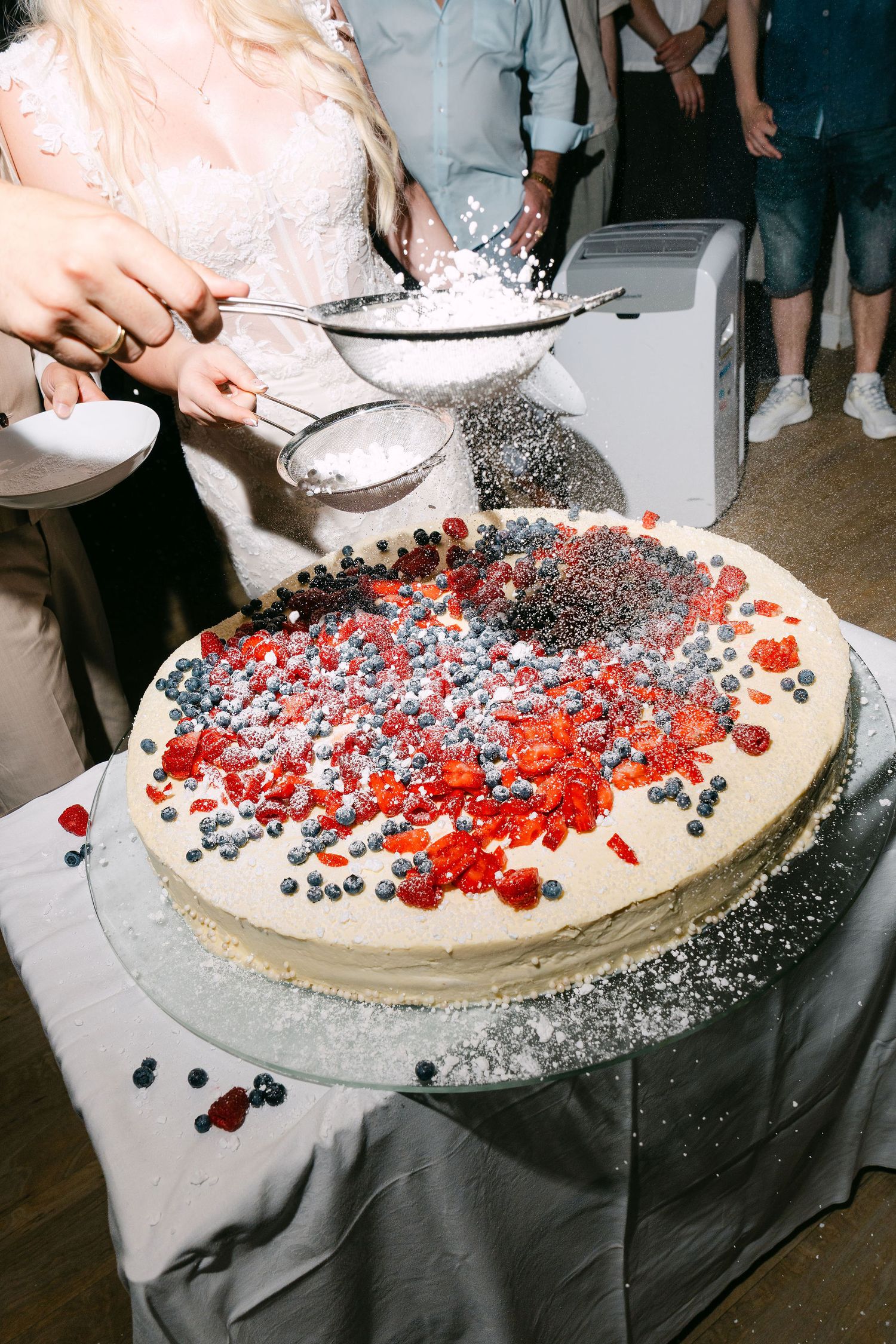Person sifting powdered sugar over a large round wedding cake topped with fresh blueberries and red berries.