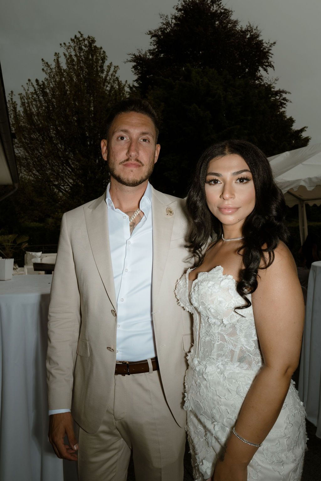A couple poses at a wedding reception, with greenery and white tents in the background.