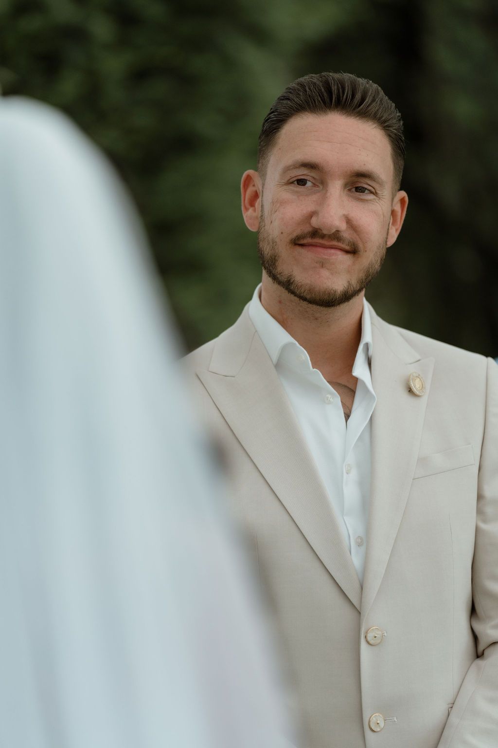 A groom in a cream-colored suit and white shirt smiles warmly while looking at someone partially visible in the foreground.