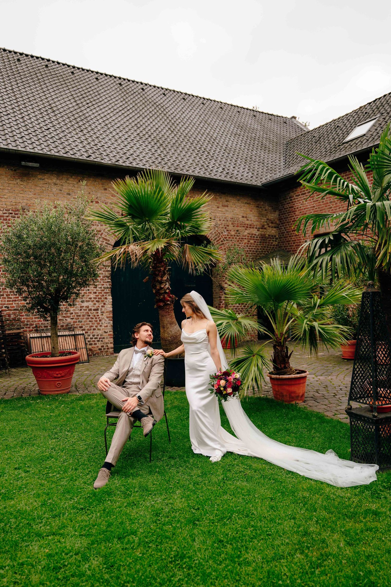 Bride in white gown and groom in beige suit pose in a courtyard garden with palm trees and brick walls.