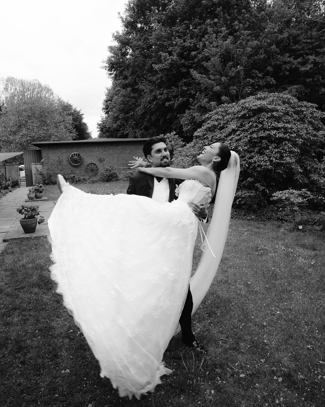 Groom lifts bride in white wedding gown outdoors in a romantic black and white photo.
