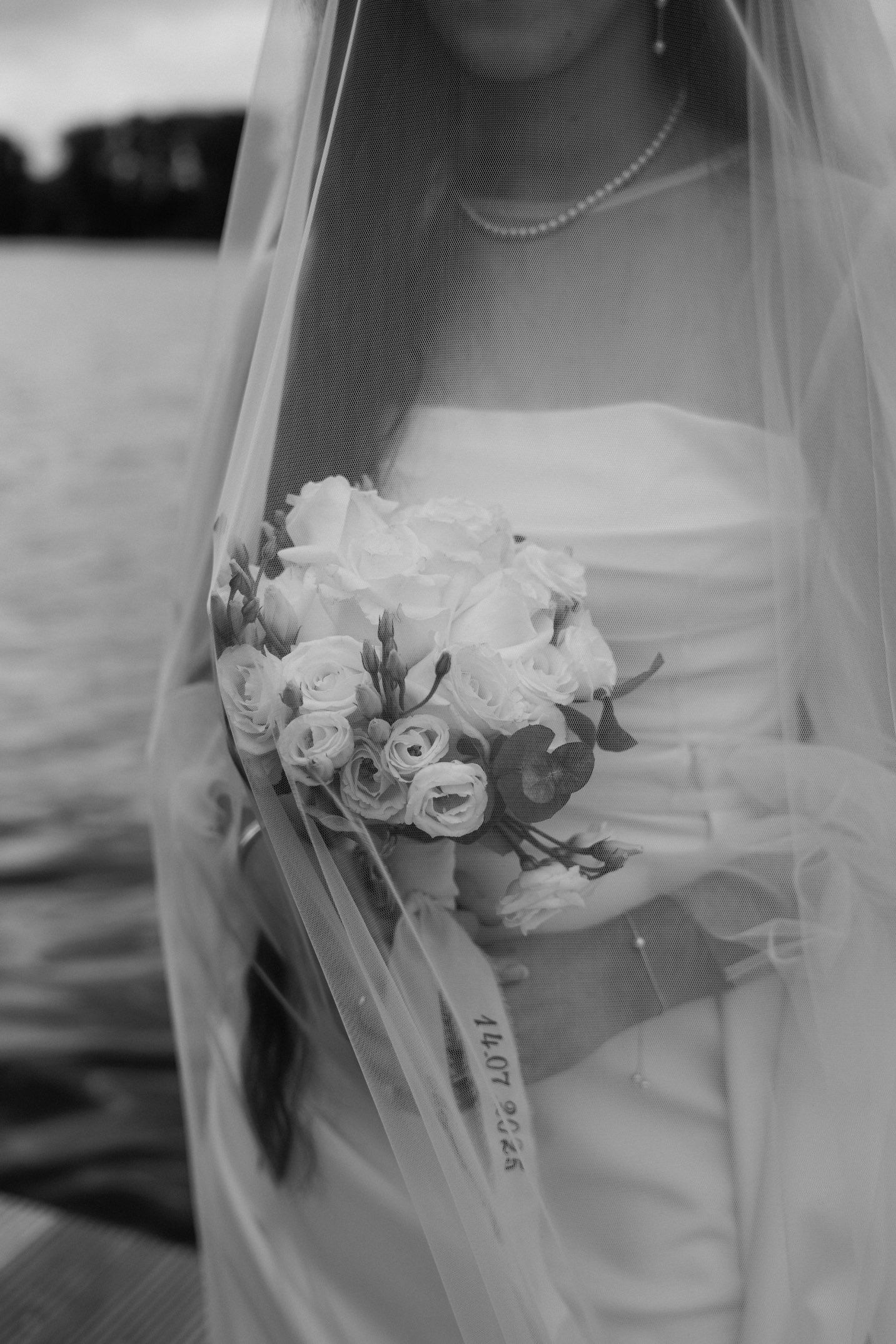 A bride holds a delicate white bridal bouquet with roses and wispy tulle veil in black and white photography.