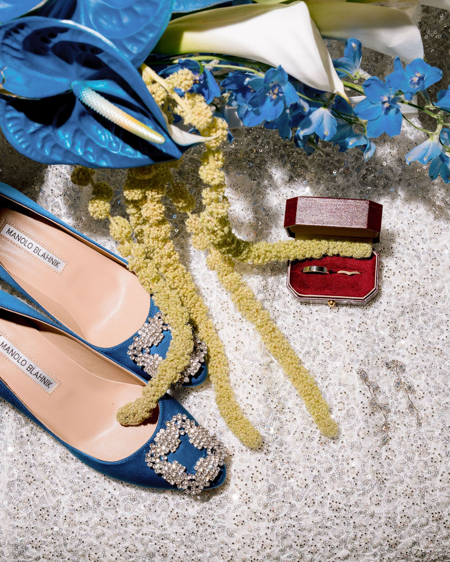 Blue Manolo Blahnik heels with crystal embellishment beside a red ring box and wedding rings on a marble surface.