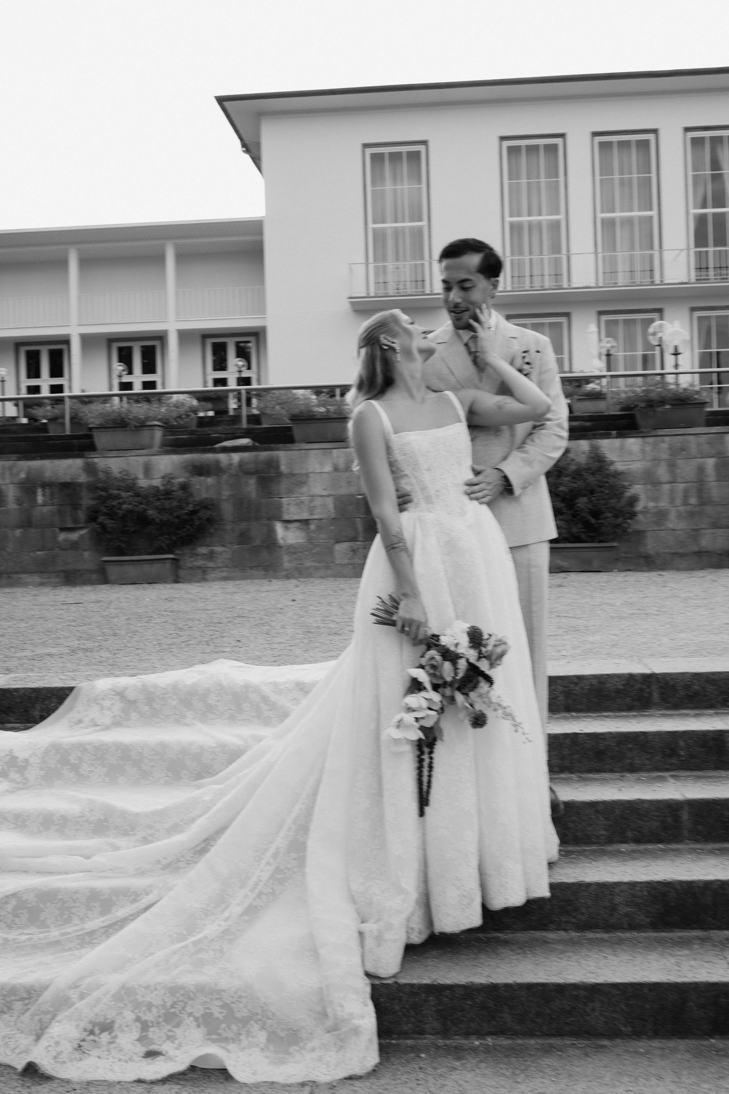 Bride and groom pose on stone steps outside elegant venue in classic black and white wedding photo.