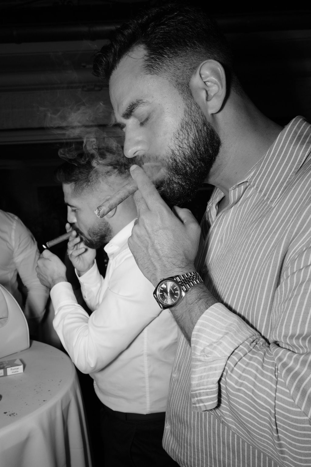 Two people in formal attire lighting cigars together in a black and white photo.
