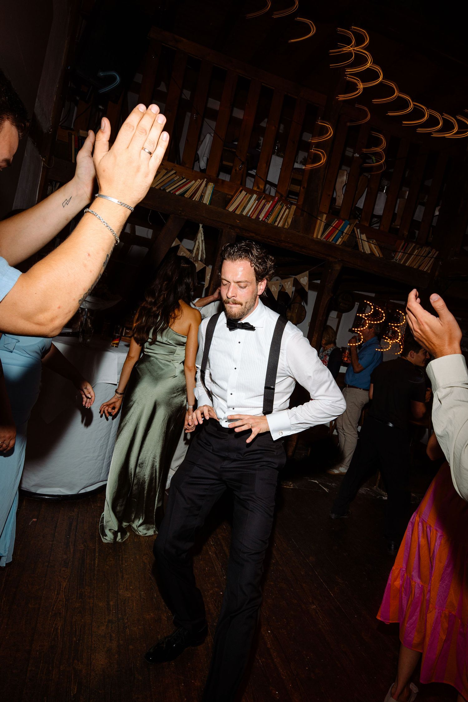 Groom in suspenders and bow tie dances energetically with guests on wedding reception dance floor.