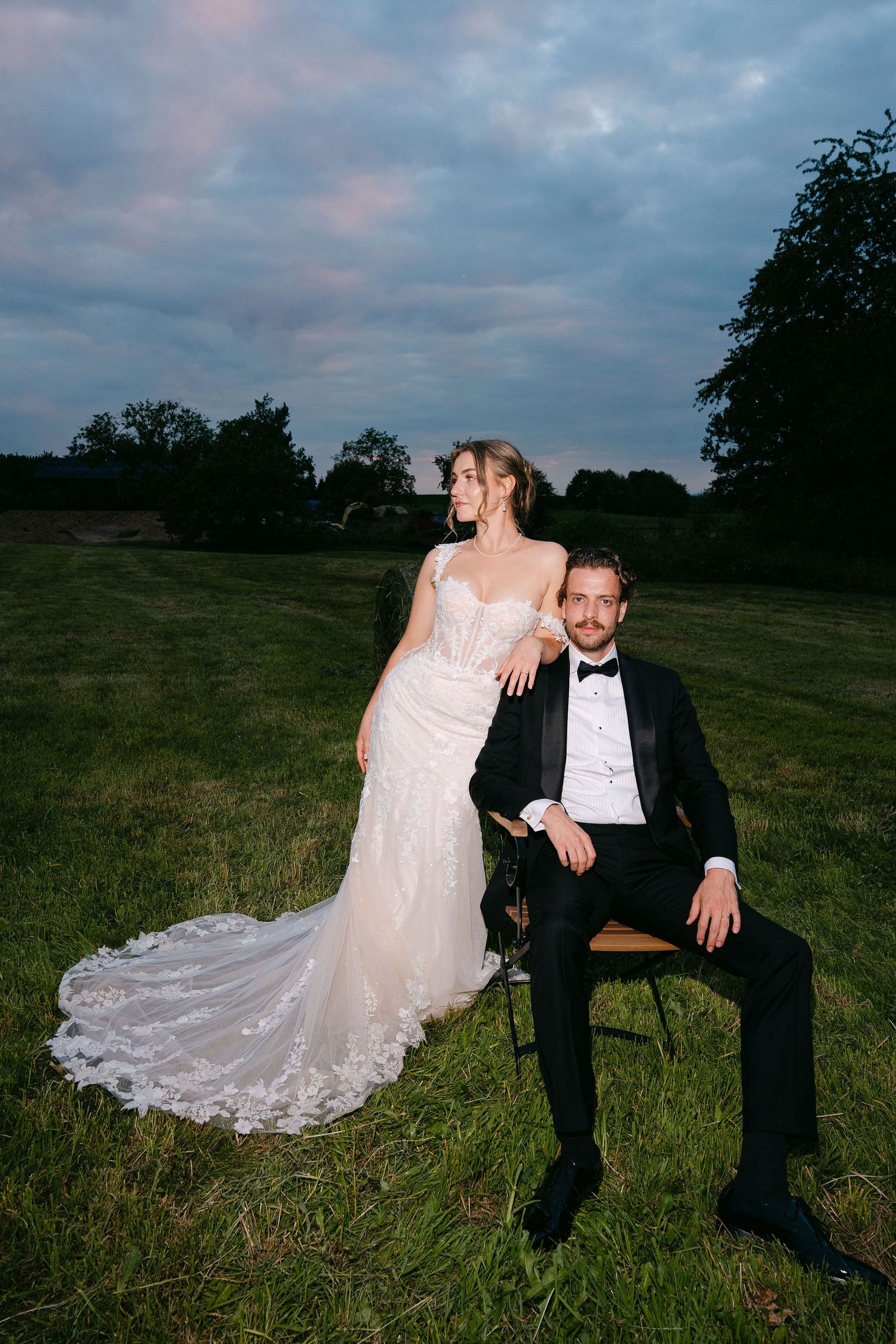 Bride in lace gown leans on seated groom in tuxedo on green field at dusk with dramatic sky.
