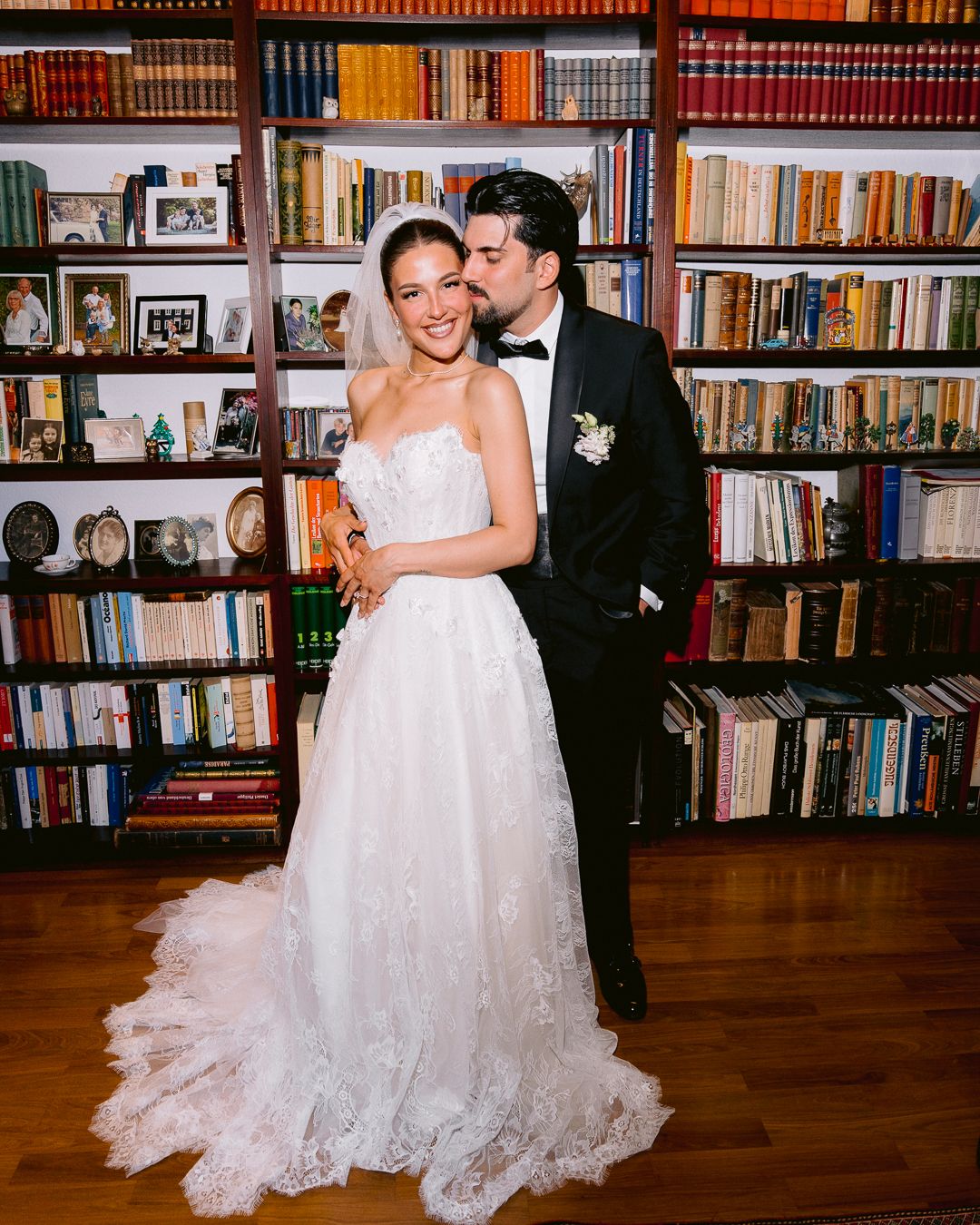 Newlyweds embrace in front of a bookshelf, bride in lace gown as groom in tuxedo kisses her cheek.