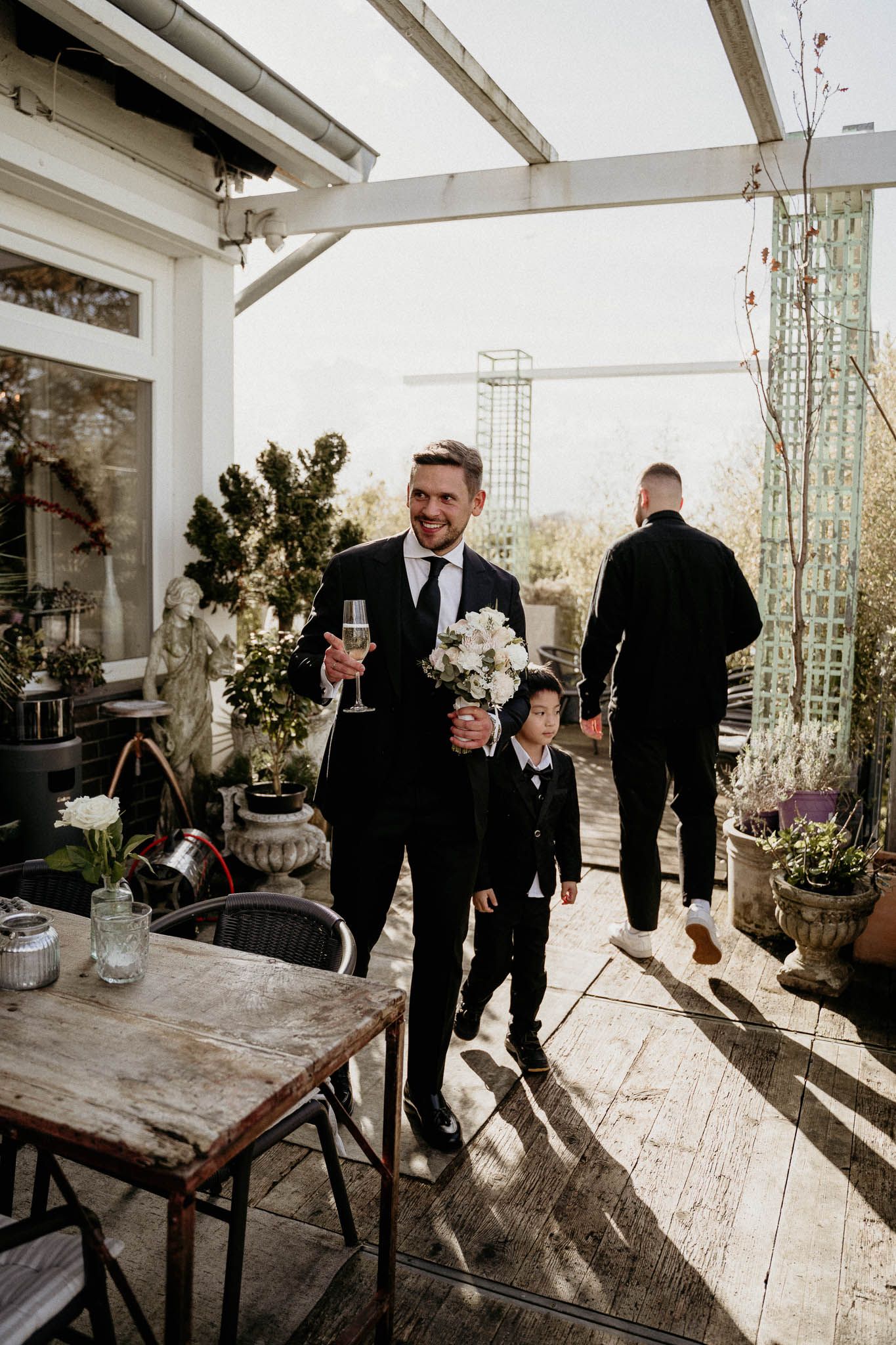 Person in a black suit holding champagne stands with a child on a rustic outdoor deck with white beams overhead.