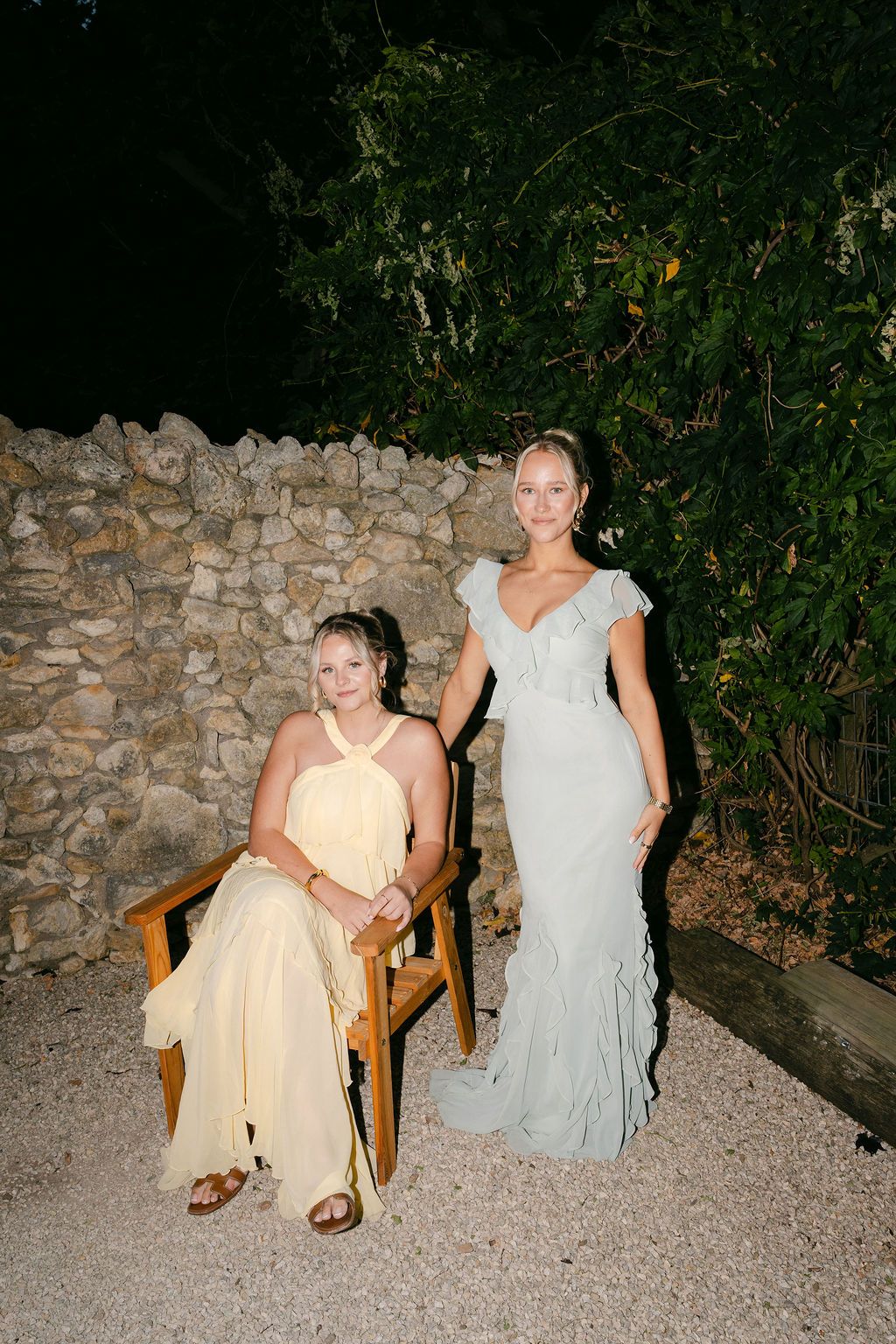 People in light colored dresses sitting on wooden chairs against a stone wall at dusk.