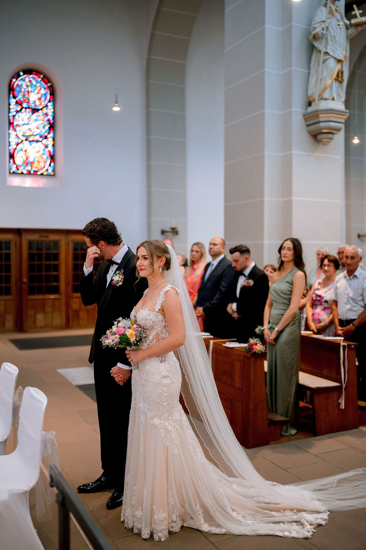 Emotional groom wipes tears beside bride in lace mermaid gown during church wedding ceremony.